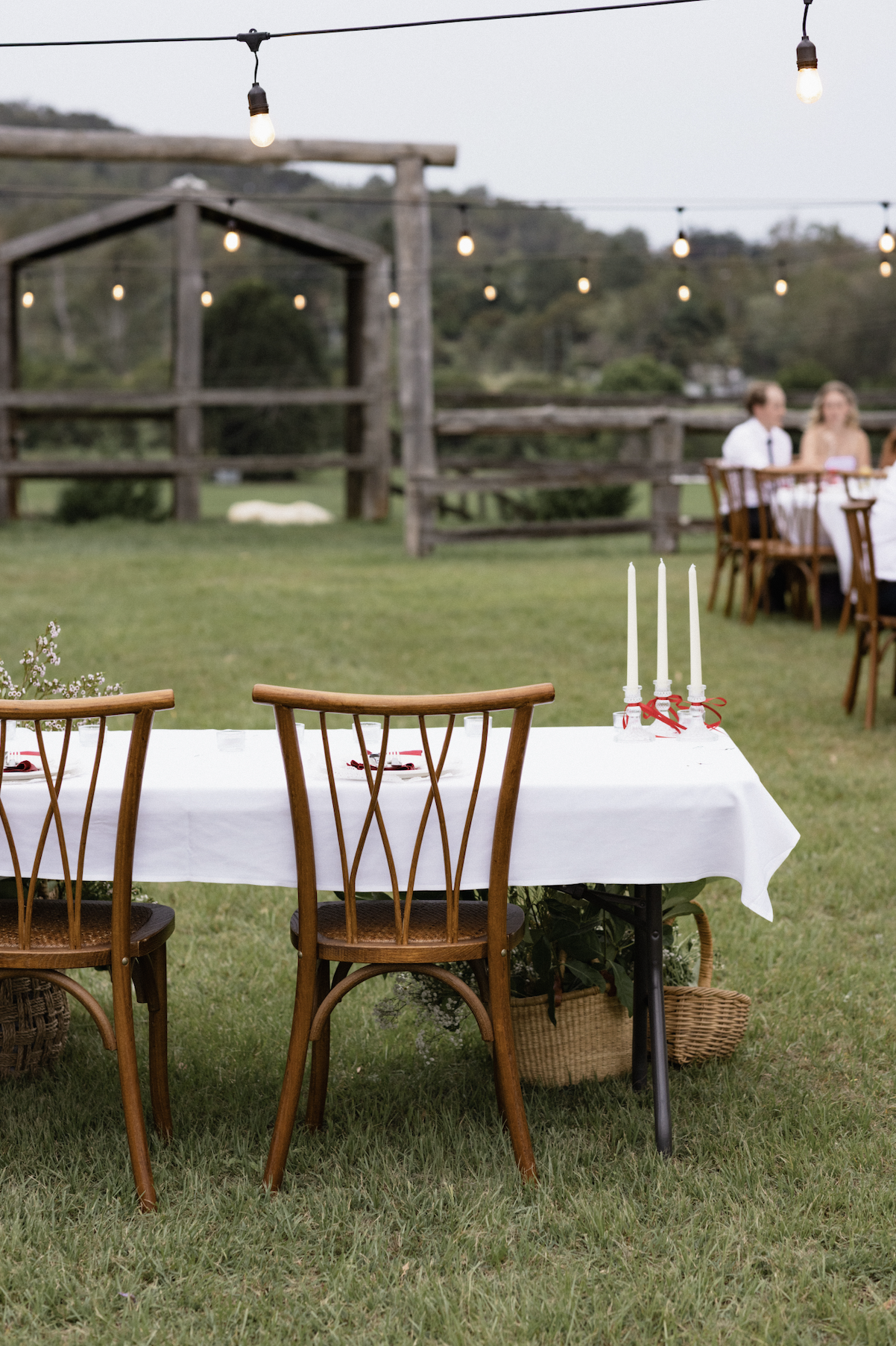 Outdoor wedding reception setup with a long white tablecloth-covered table, three white taper candles with red ribbons, wooden chairs, and string lights hanging in a grassy field with a rustic wooden fence and distant trees.