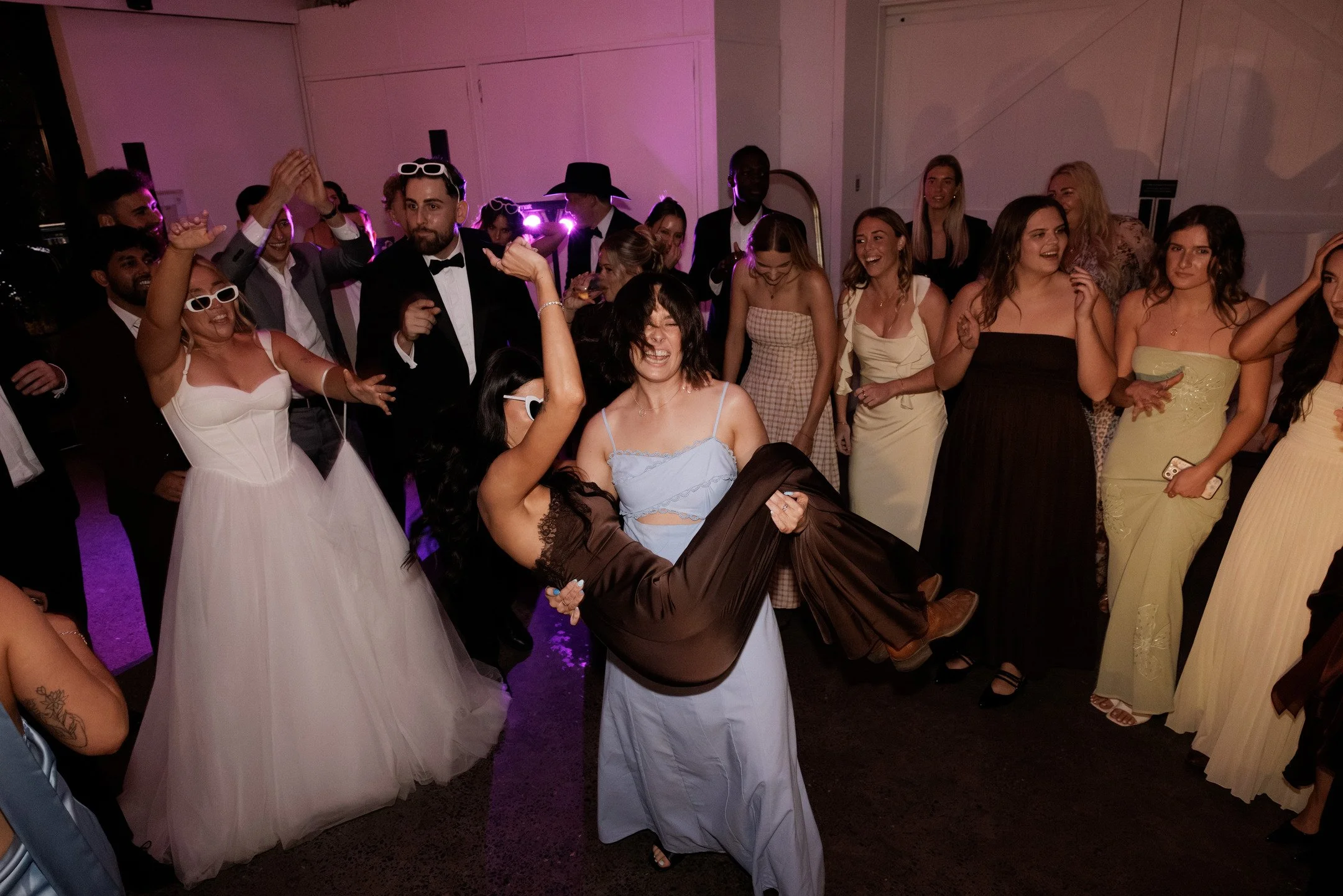 People dancing and celebrating at a party, with some wearing formal and party attire, in a dimly lit room with purple lighting.