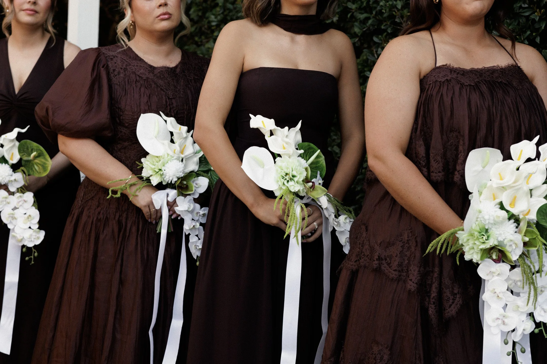 Women dressed in dark brown dresses holding white floral bouquets at an outdoor event.