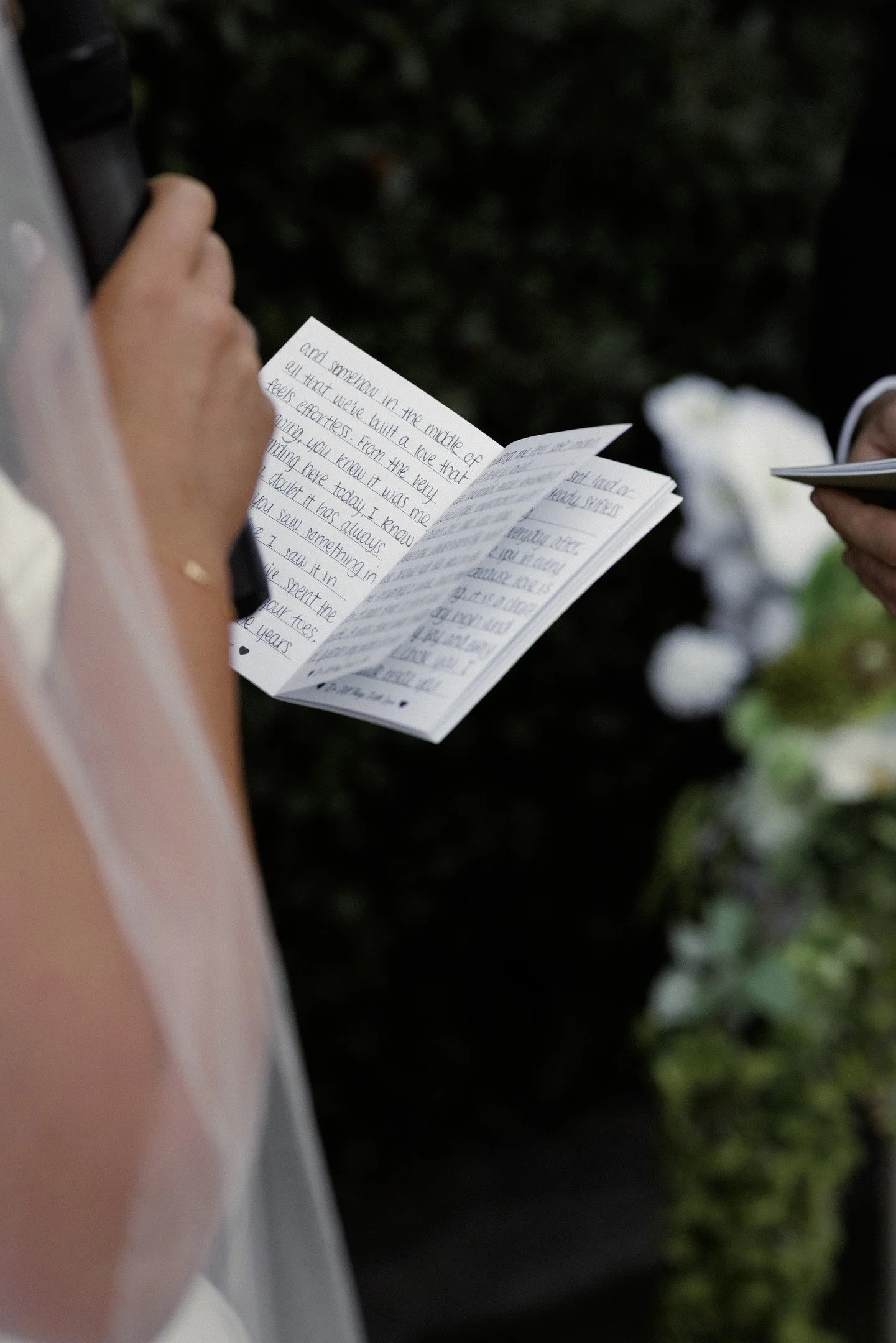A person holding a folded piece of paper with handwritten notes, outdoors at night, with another person visible in the background.
