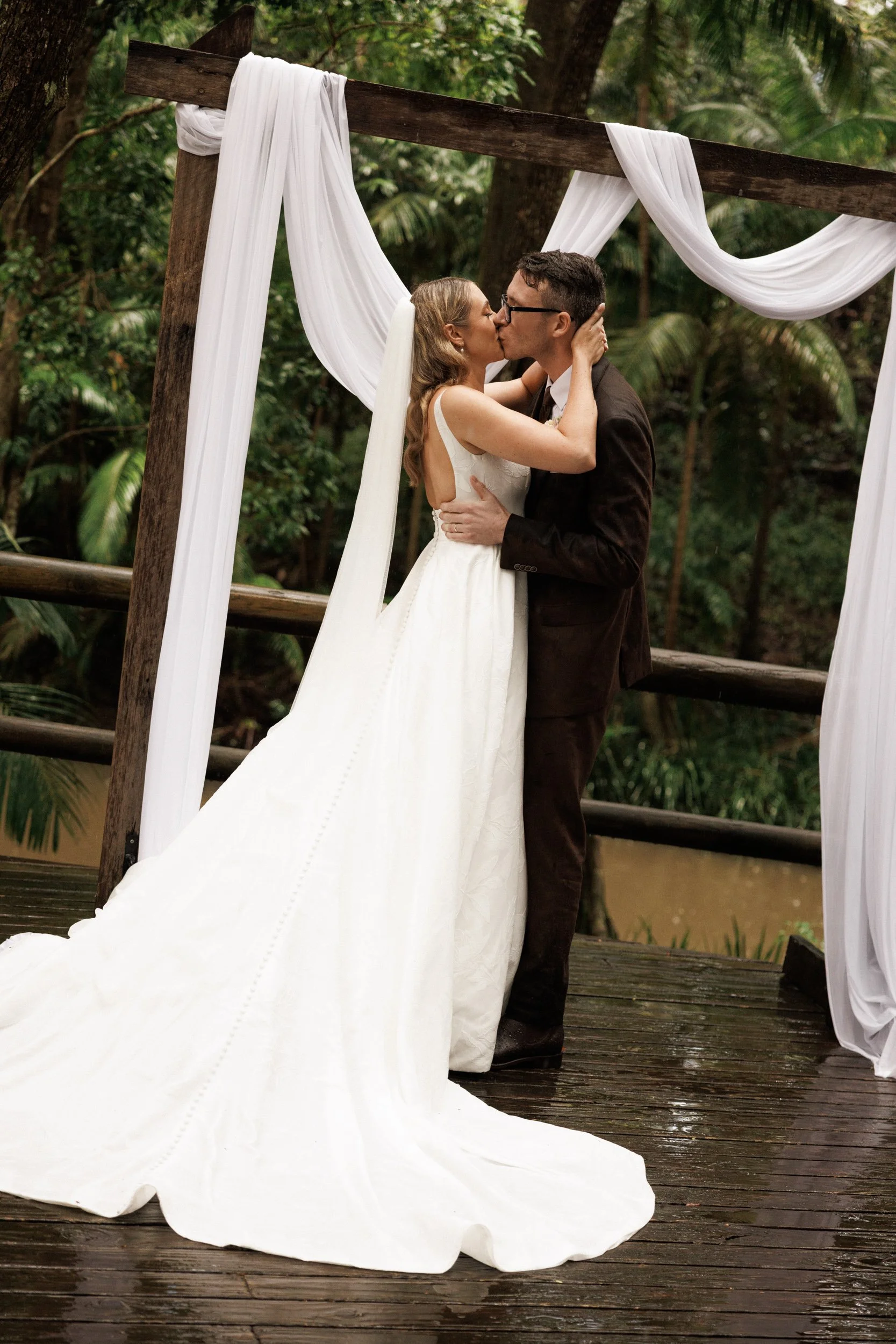 A couple kissing under a wooden arch decorated with white drapes in an outdoor setting with trees and greenery.