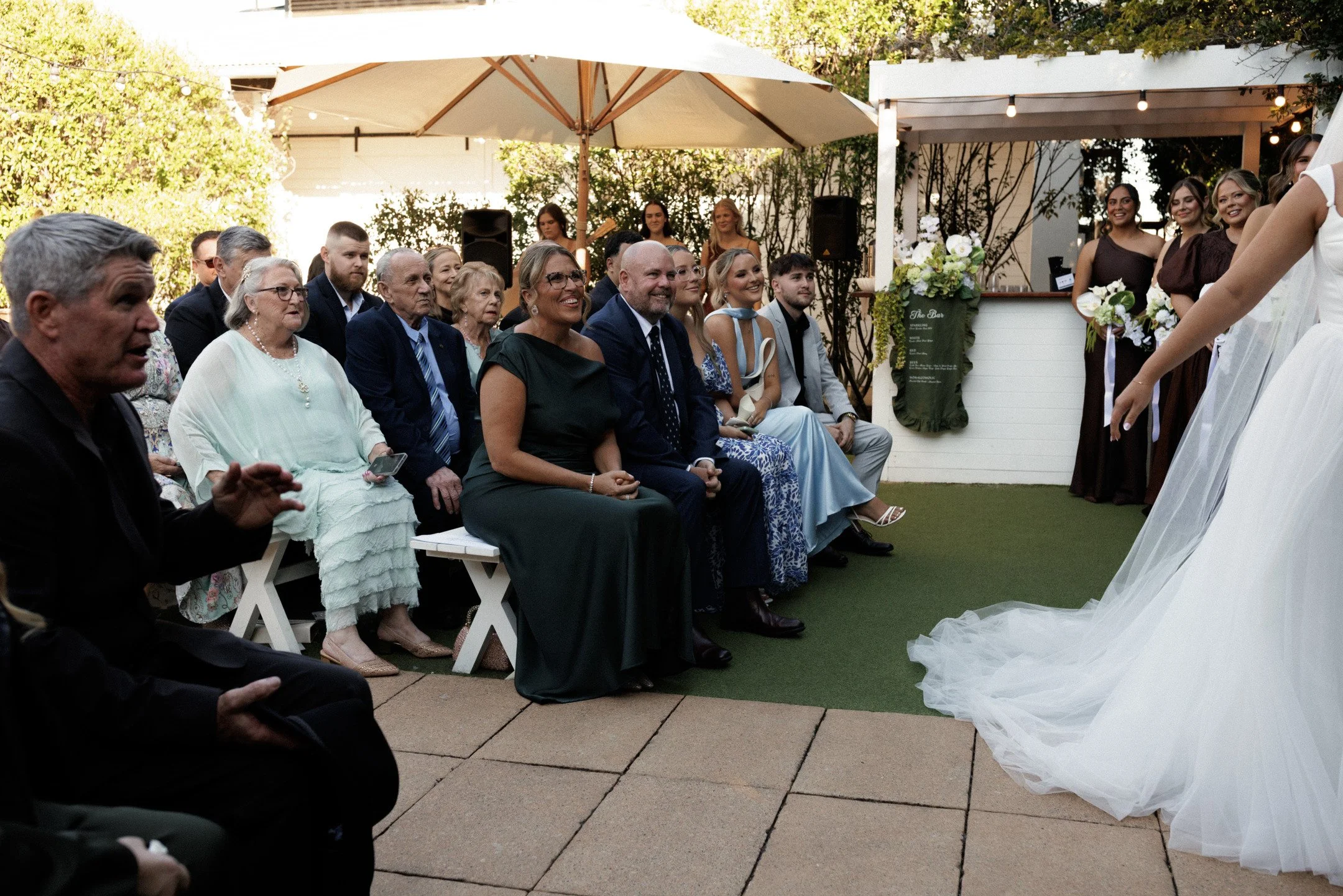 Wedding ceremony outdoors with seated guests and wedding dress visible in the foreground.