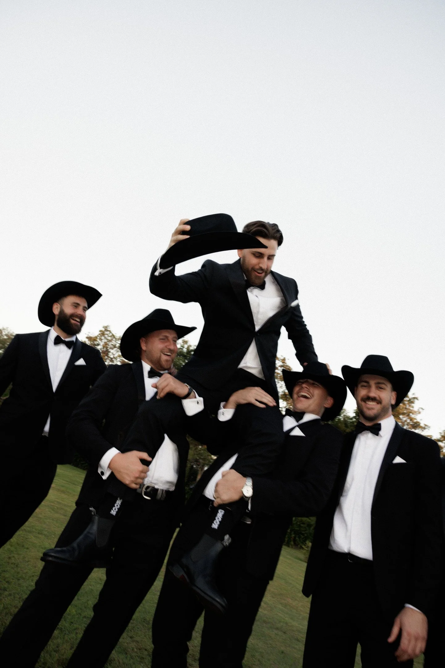 Group of men in tuxedos and cowboy hats celebrating outdoors.