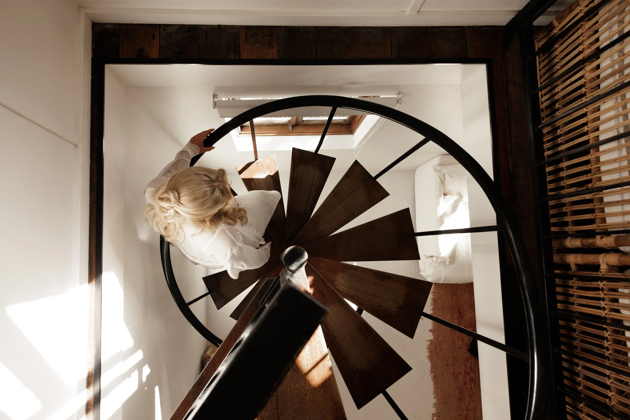 A person with blonde curly hair, dressed in white, seen from above, ascending a spiral staircase with dark wooden steps and a black metal railing, in a well-lit indoor space.