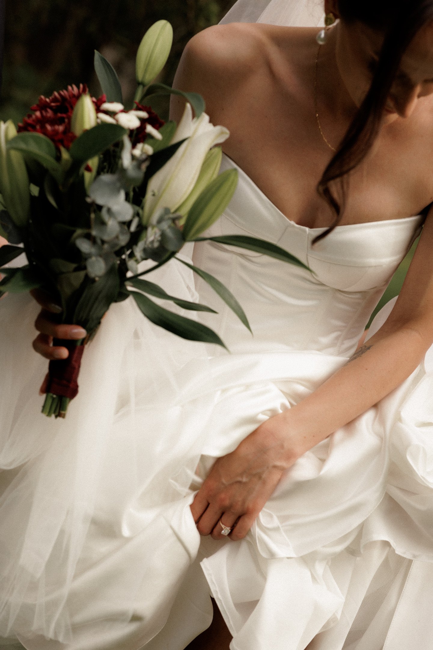 A bride in a white strapless wedding gown sitting on a white surface, holding a bouquet of white lilies, red flowers, and greenery, with her left hand resting on her lap showing a wedding ring.