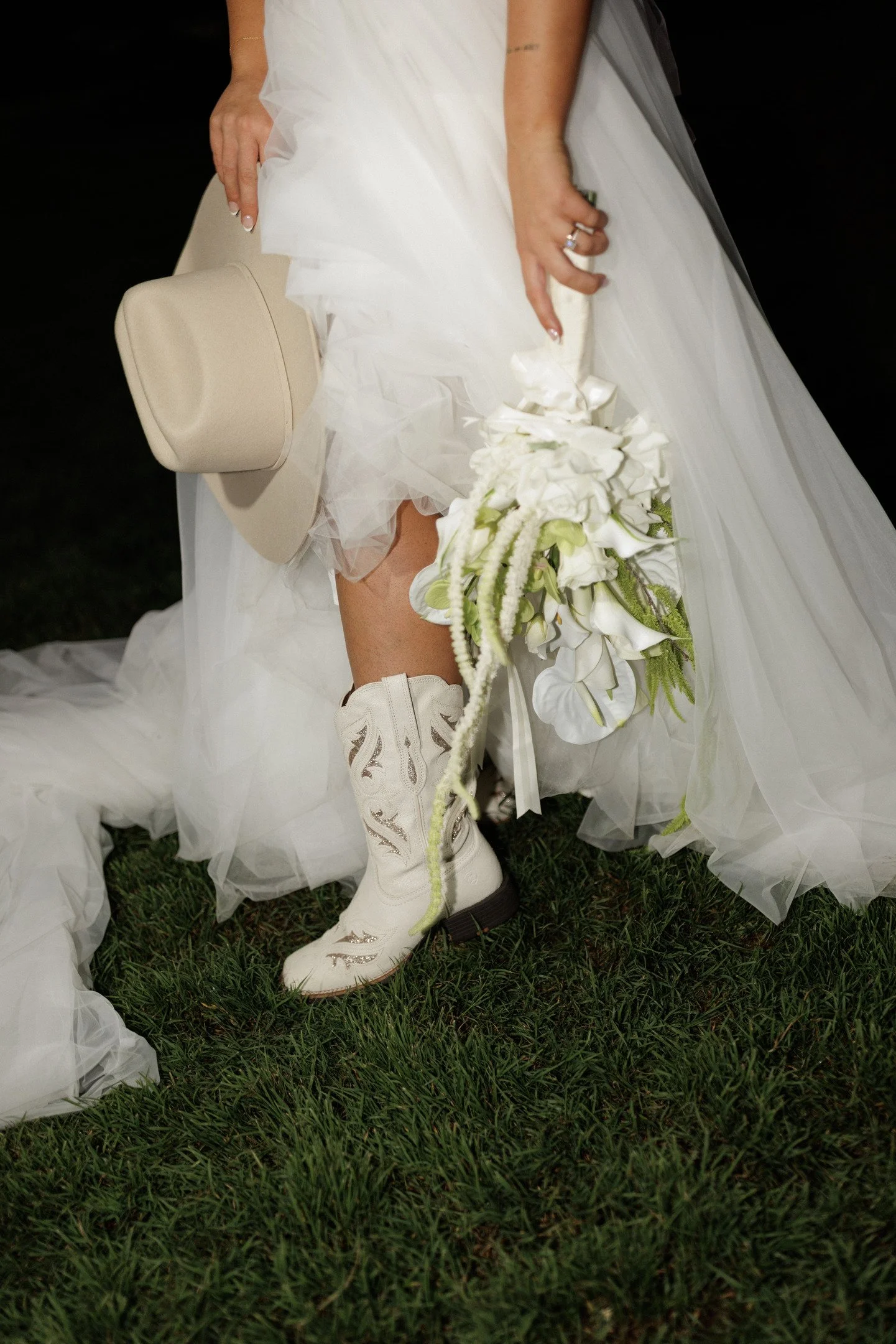 Close-up of a person dressed in white cowboy boots and a flowing white dress, holding a beige hat and a bouquet of white flowers, standing on grass.