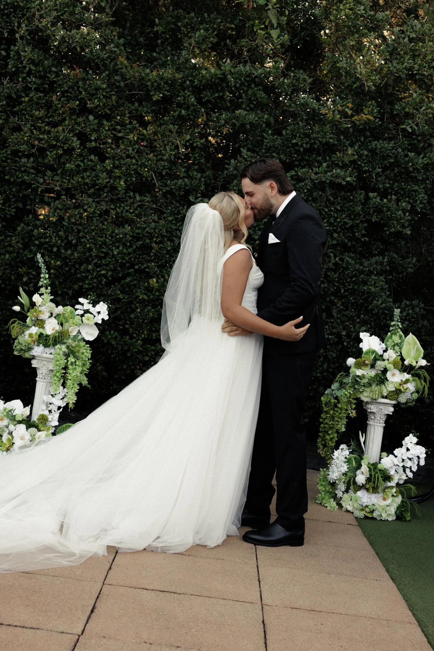A bride and groom kissing during their wedding ceremony outdoors, with greenery and floral decorations in the background.