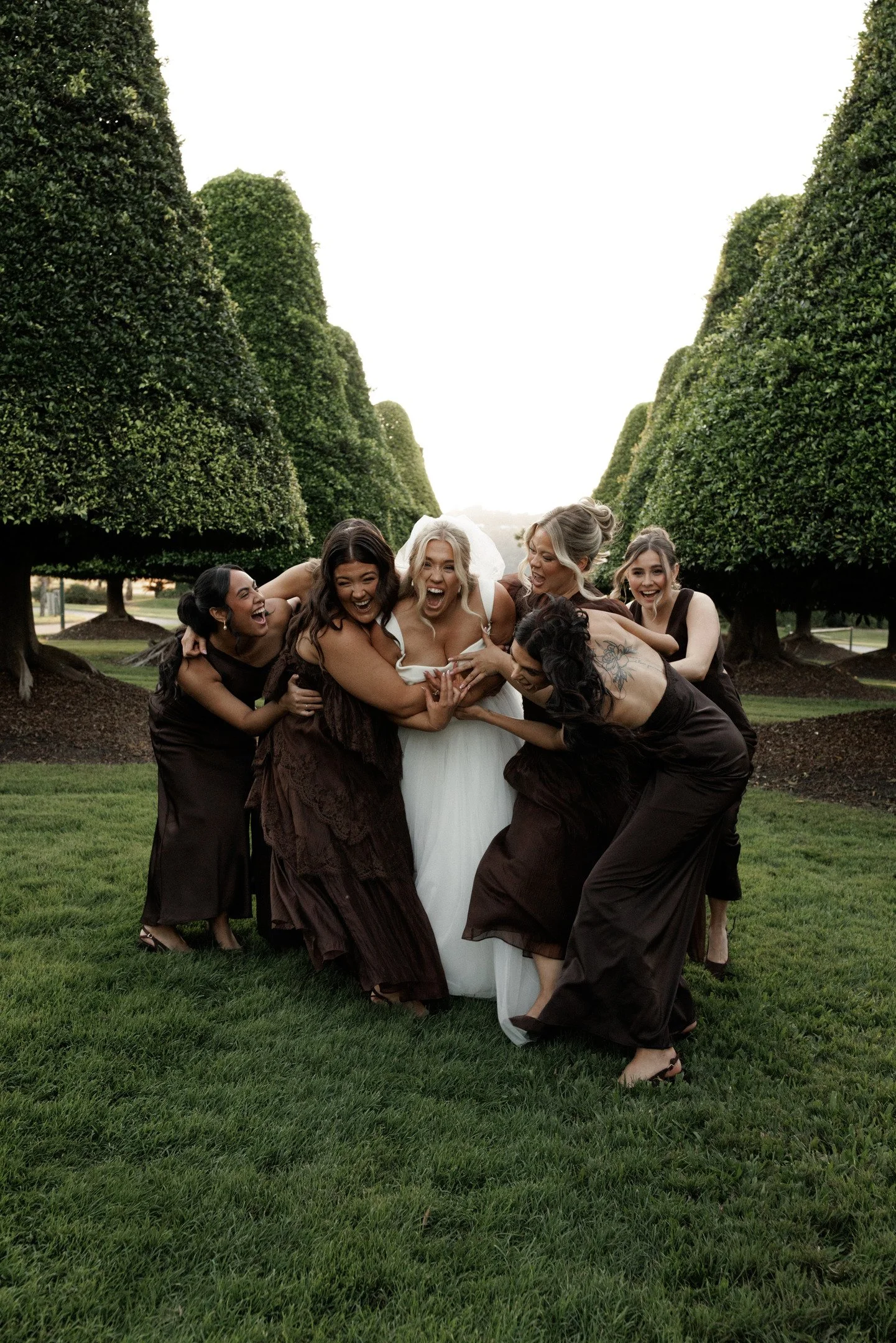 Group of women in brown dresses surrounding a woman in a white dress, outdoors on a grassy area with tall, trimmed trees in the background, all smiling and laughing.