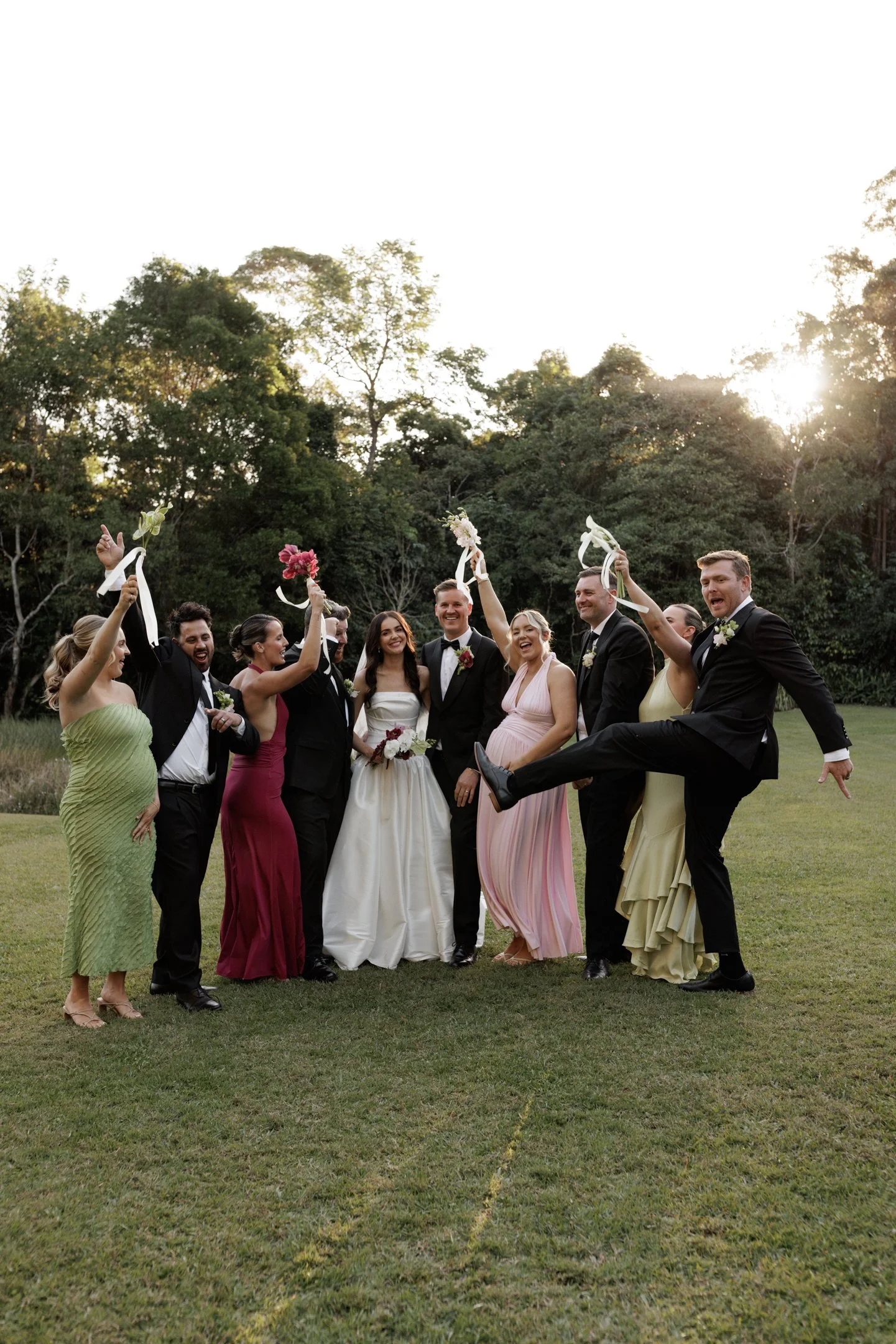 Group of men and women dressed in formal attire celebrating at a wedding outdoors, with trees and sunlight in the background.