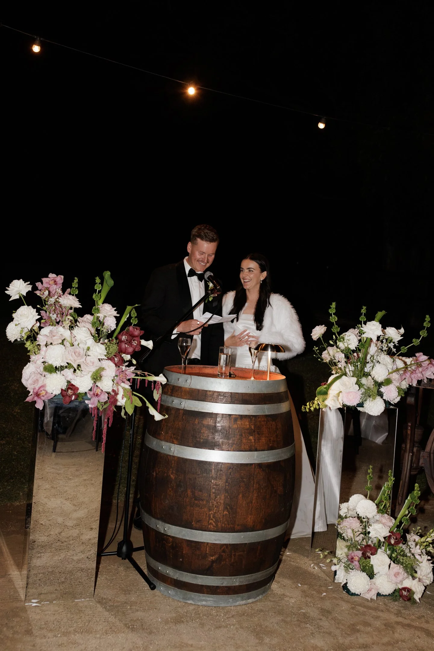 A man and woman stand together smiling during a wedding ceremony at night, with floral arrangements and a barrel table in front of them, under string lights.