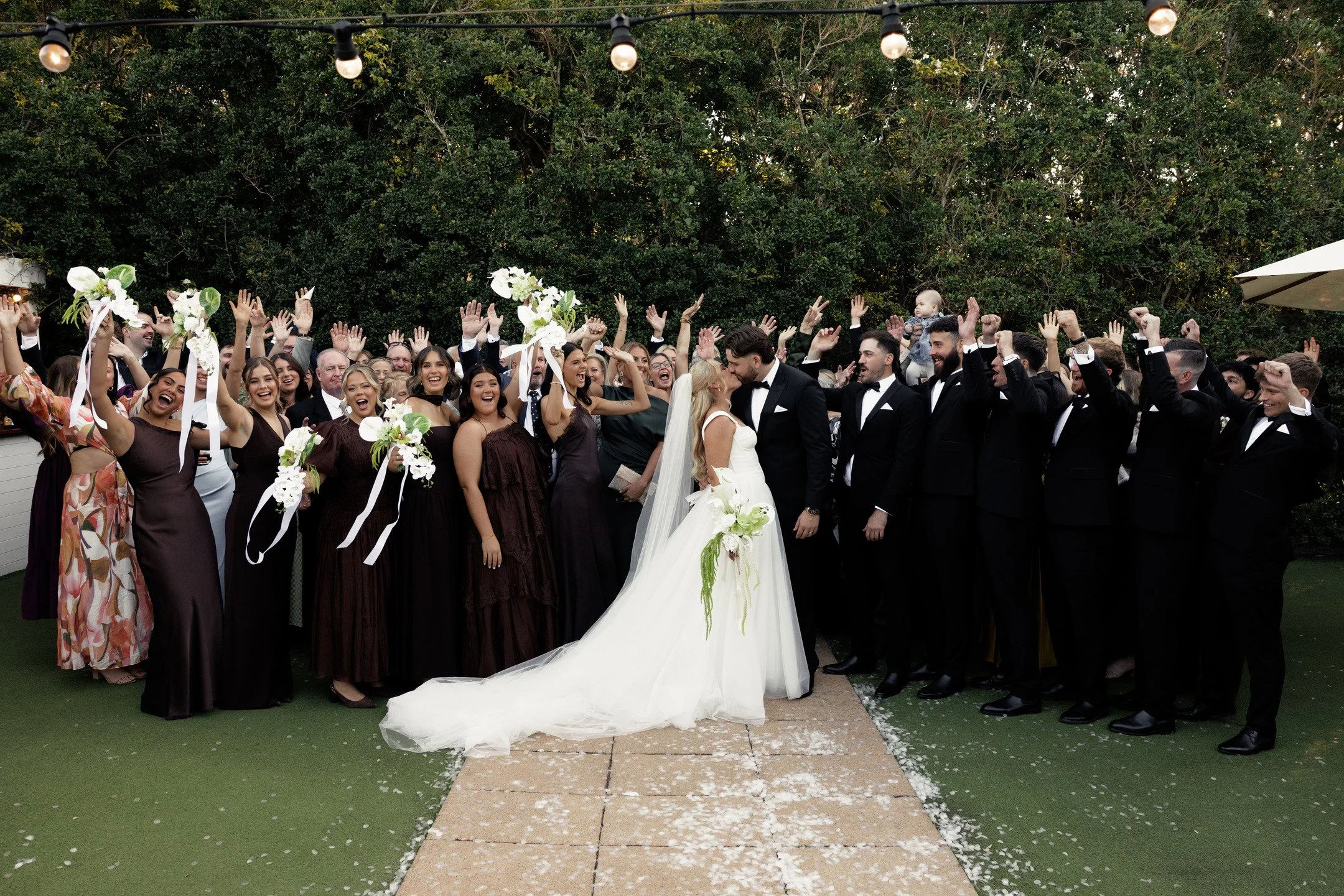 A wedding celebration with a large group of guests gathered around the bride and groom, who are sharing a kiss, outdoors under string lights and trees.