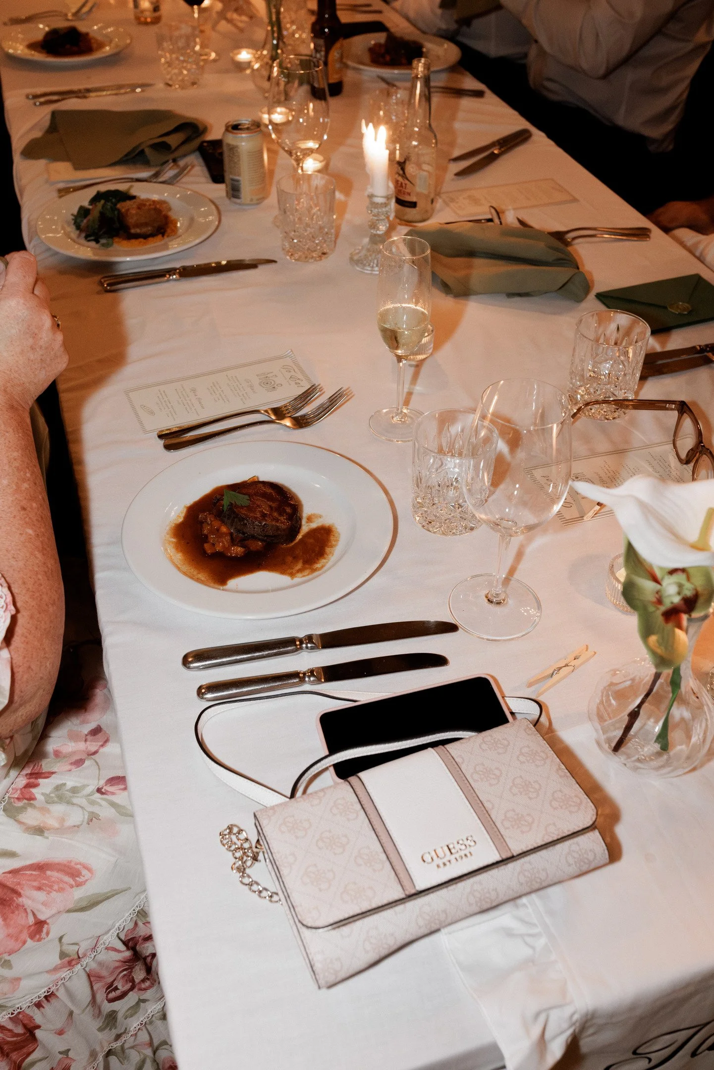 A dinner table set with plates of food, wine glasses, silverware, candles, and beverages. A purse and a Guess-themed gift box are on the table.