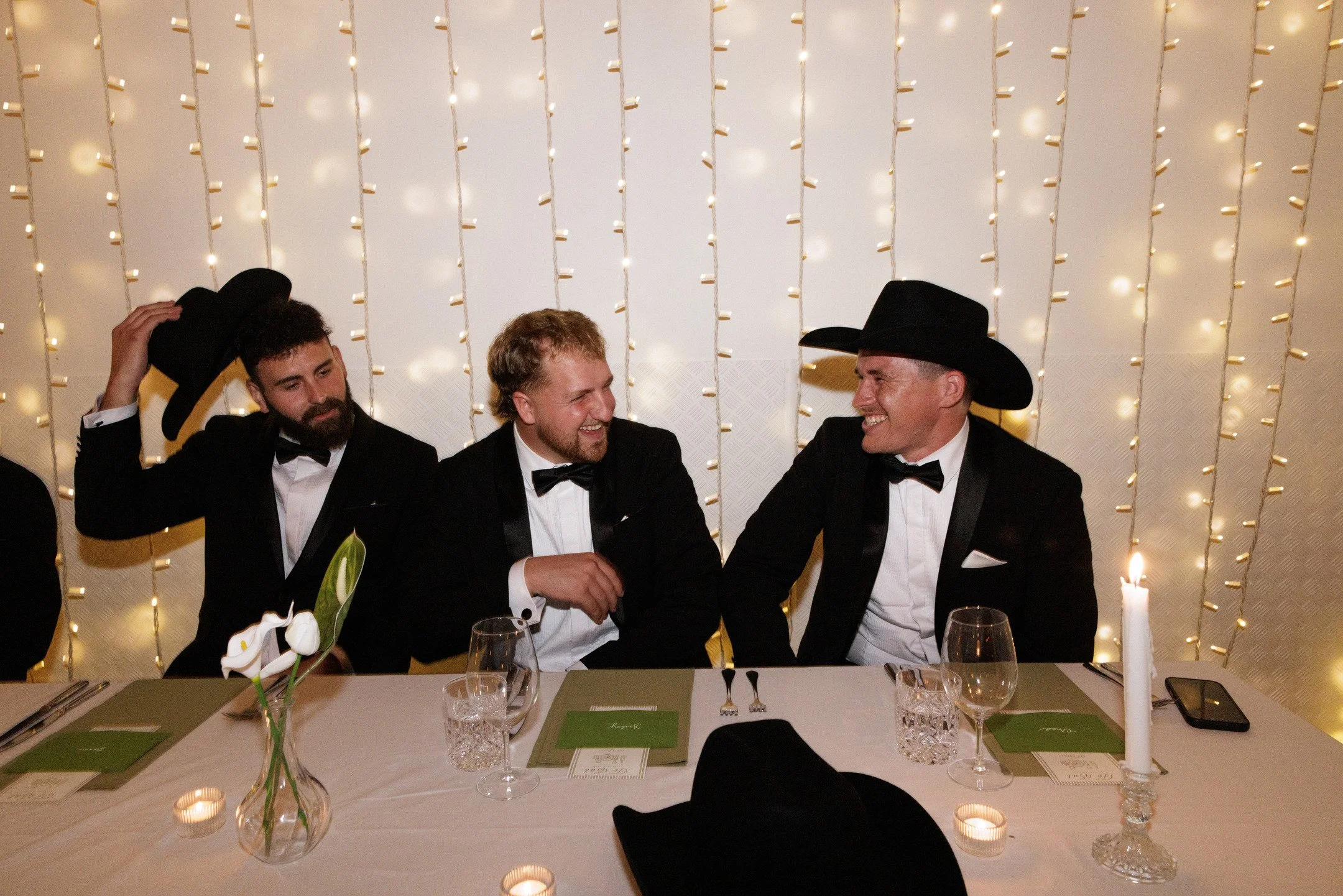 Three men in tuxedos sitting at a decorated dinner table, two wearing black cowboy hats, engaged in conversation, with floral arrangements, candles, and drinks on the table, and a backdrop of white string lights.