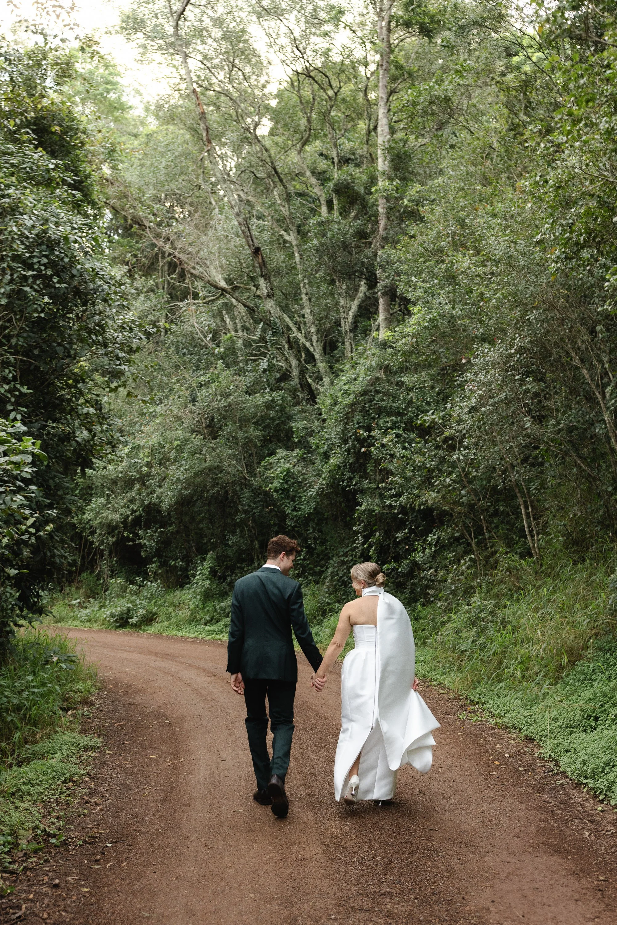 Couple in wedding attire walking on dirt path through lush forest -Elevated Weddings
