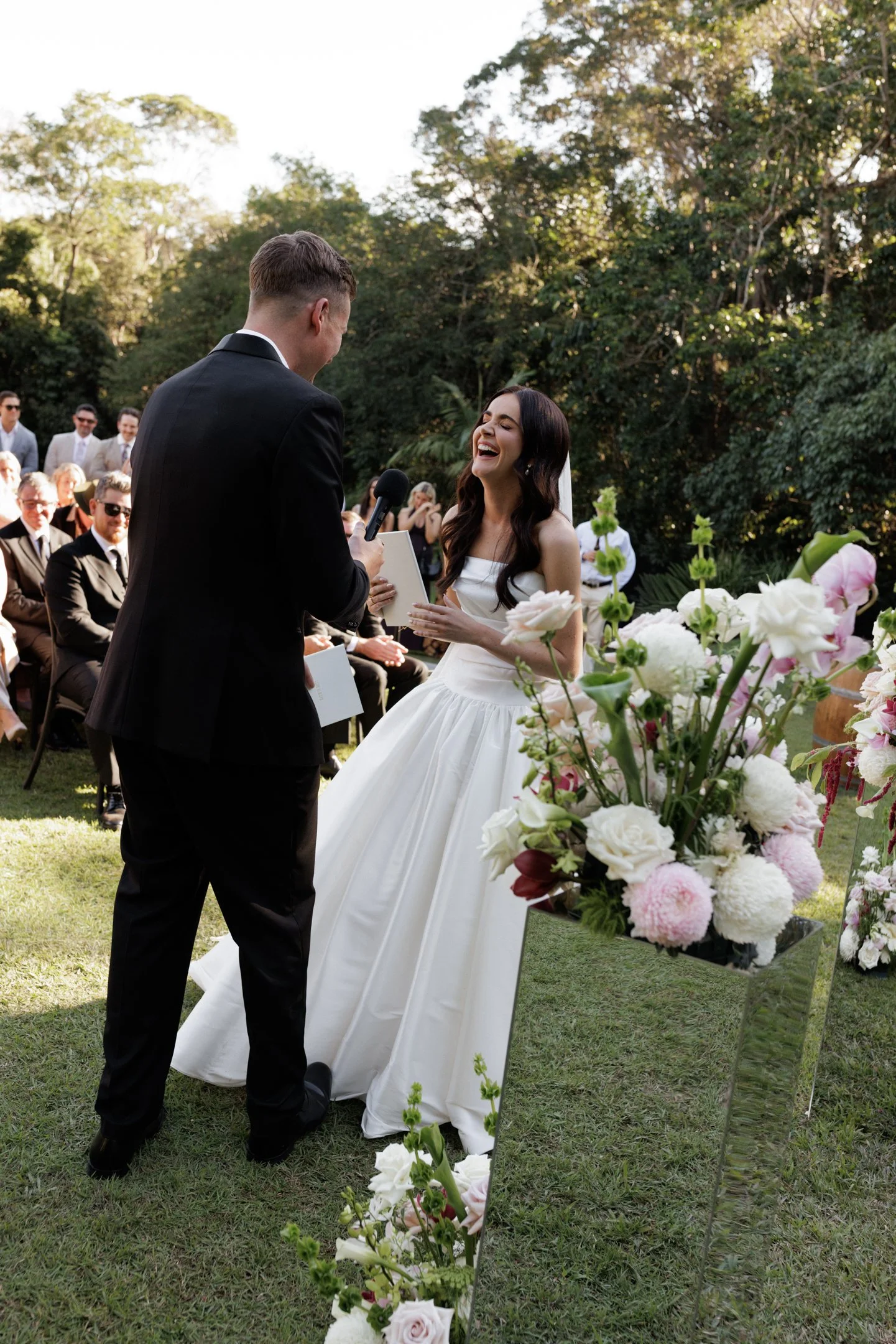 A bride and groom exchange vows at an outdoor wedding ceremony, with guests seated in the background and surrounded by lush greenery and flowers.