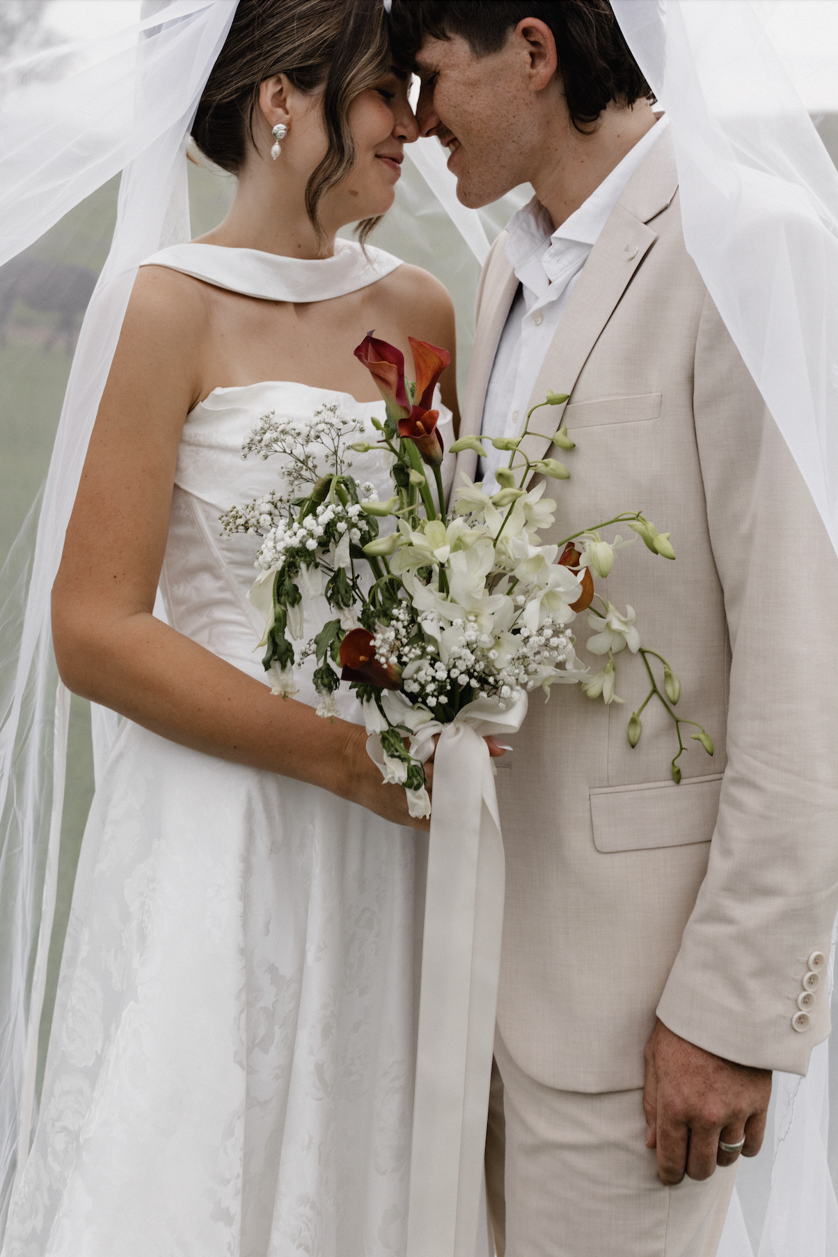 A bride and groom are touching foreheads and smiling while holding a bouquet of white and dark red flowers, with a veil draped over them, outdoors.