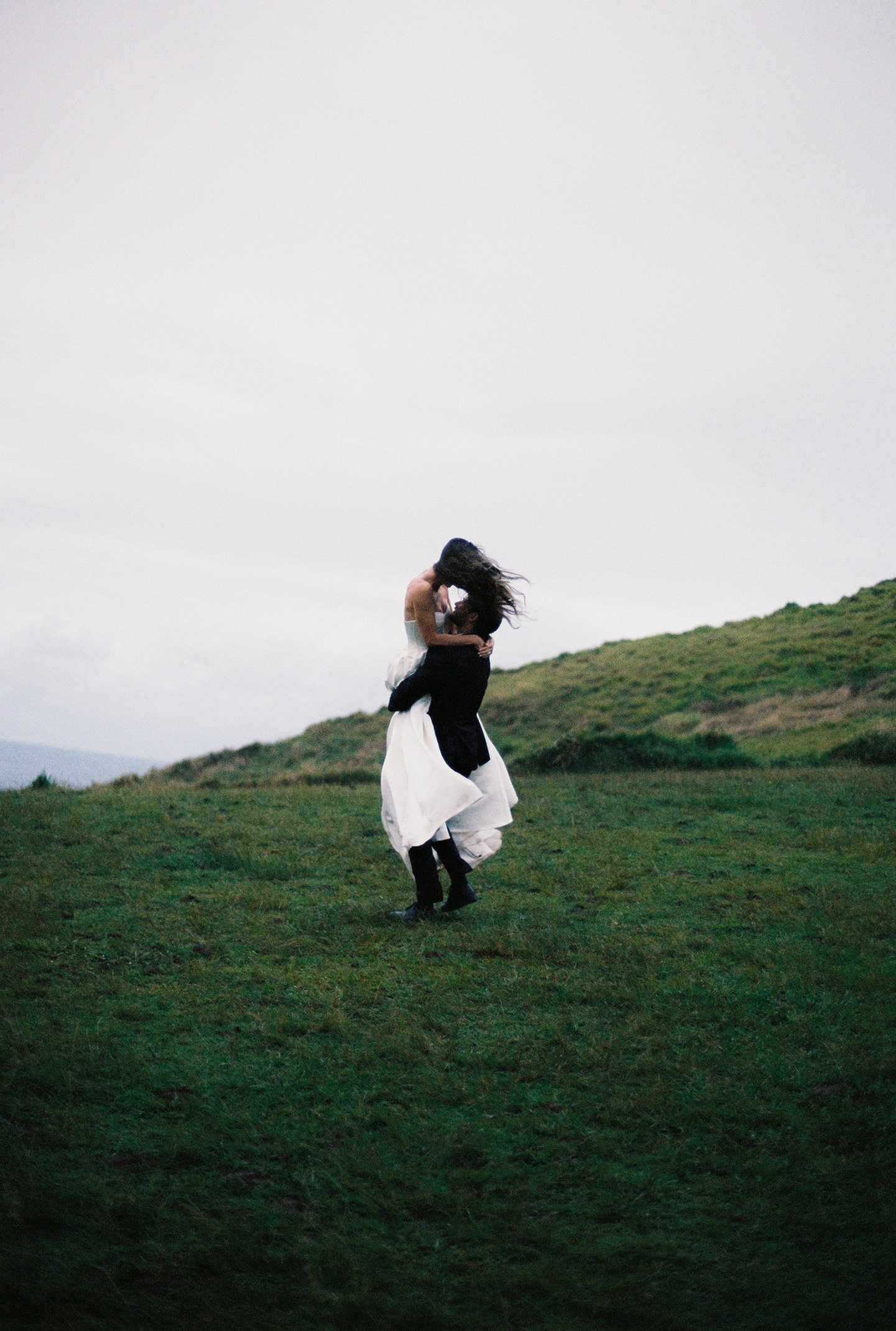 A person in a white dress is lifting another person in dark clothing onto their shoulders on a grassy hillside, with cloudy sky overhead.