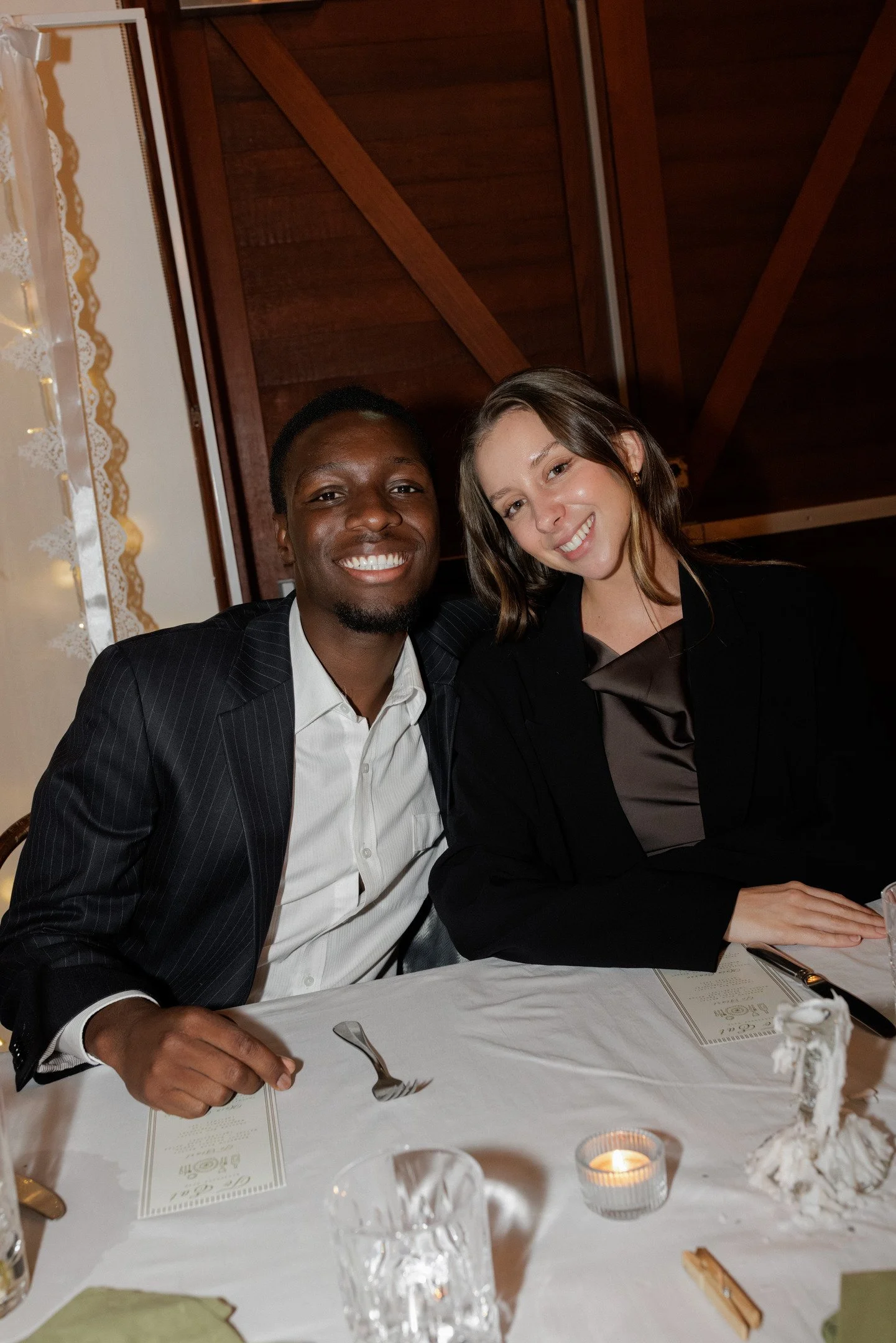 A man and a woman sitting at a dining table, smiling for a photo indoors with wooden wall behind them.