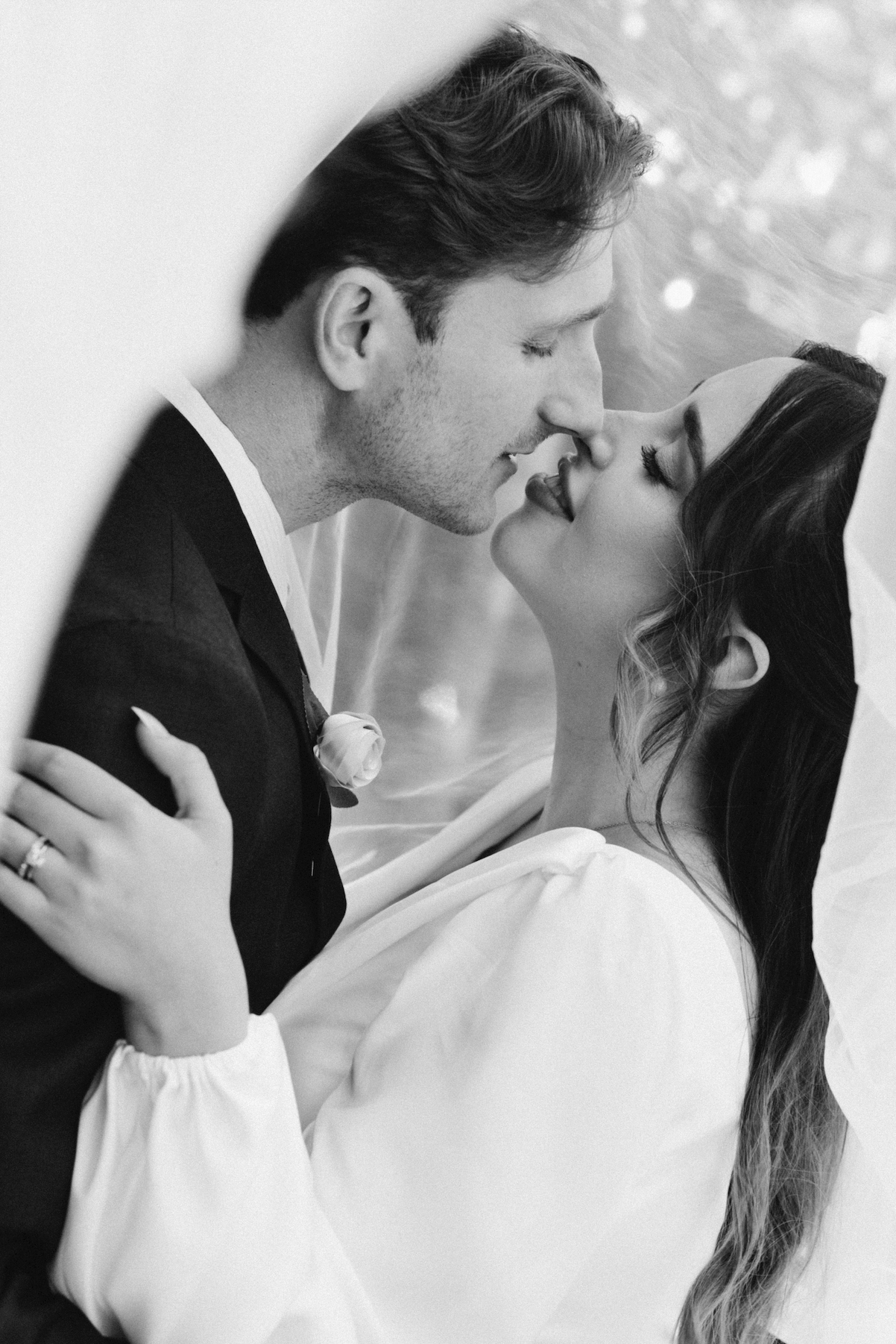 Black and white photo of a couple in wedding attire, sharing an intimate moment with their faces close, about to kiss.
