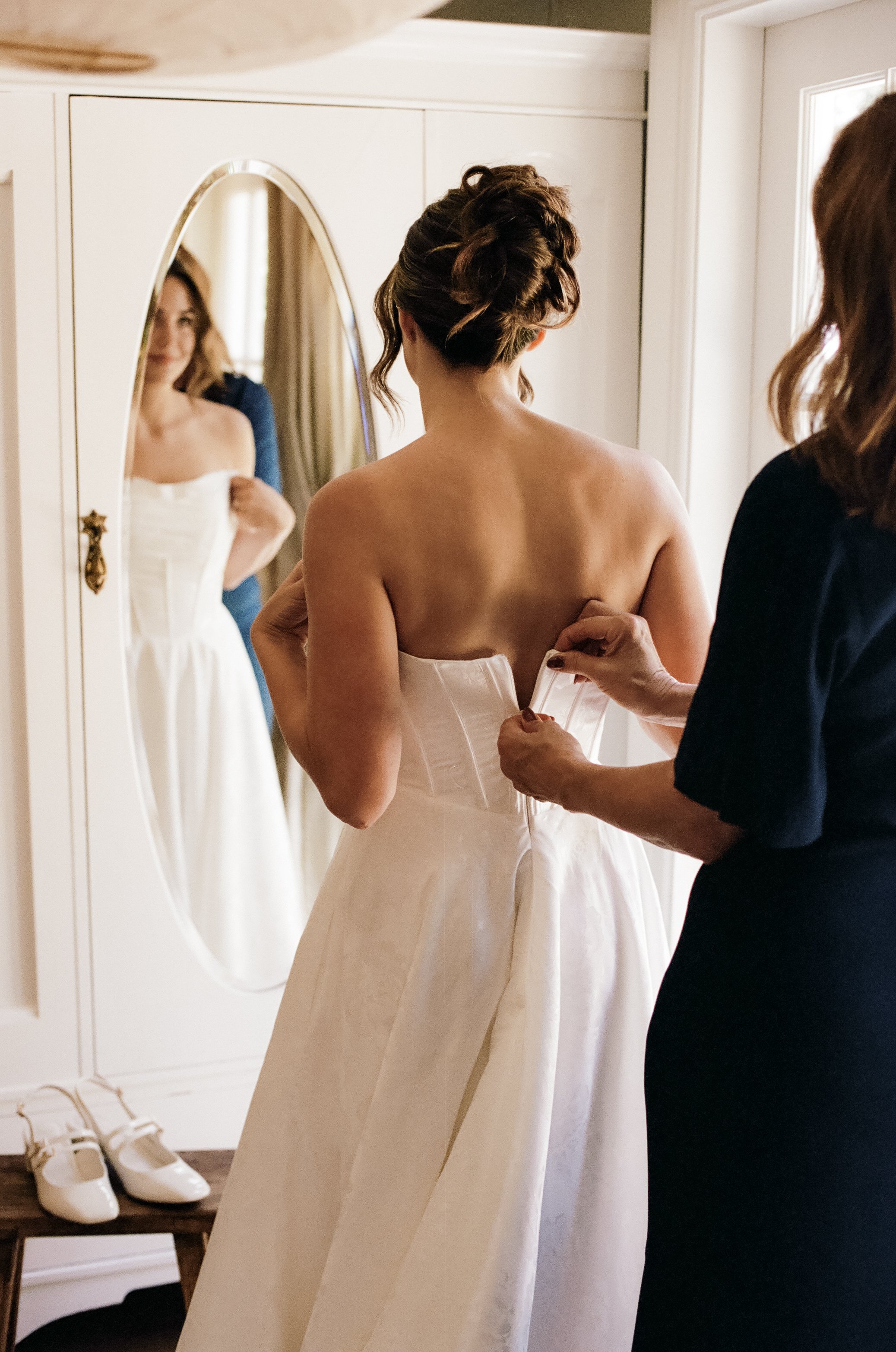 A woman in a strapless wedding dress is being helped into her gown from behind by another woman, while looking into a mirror that shows her smiling reflection.