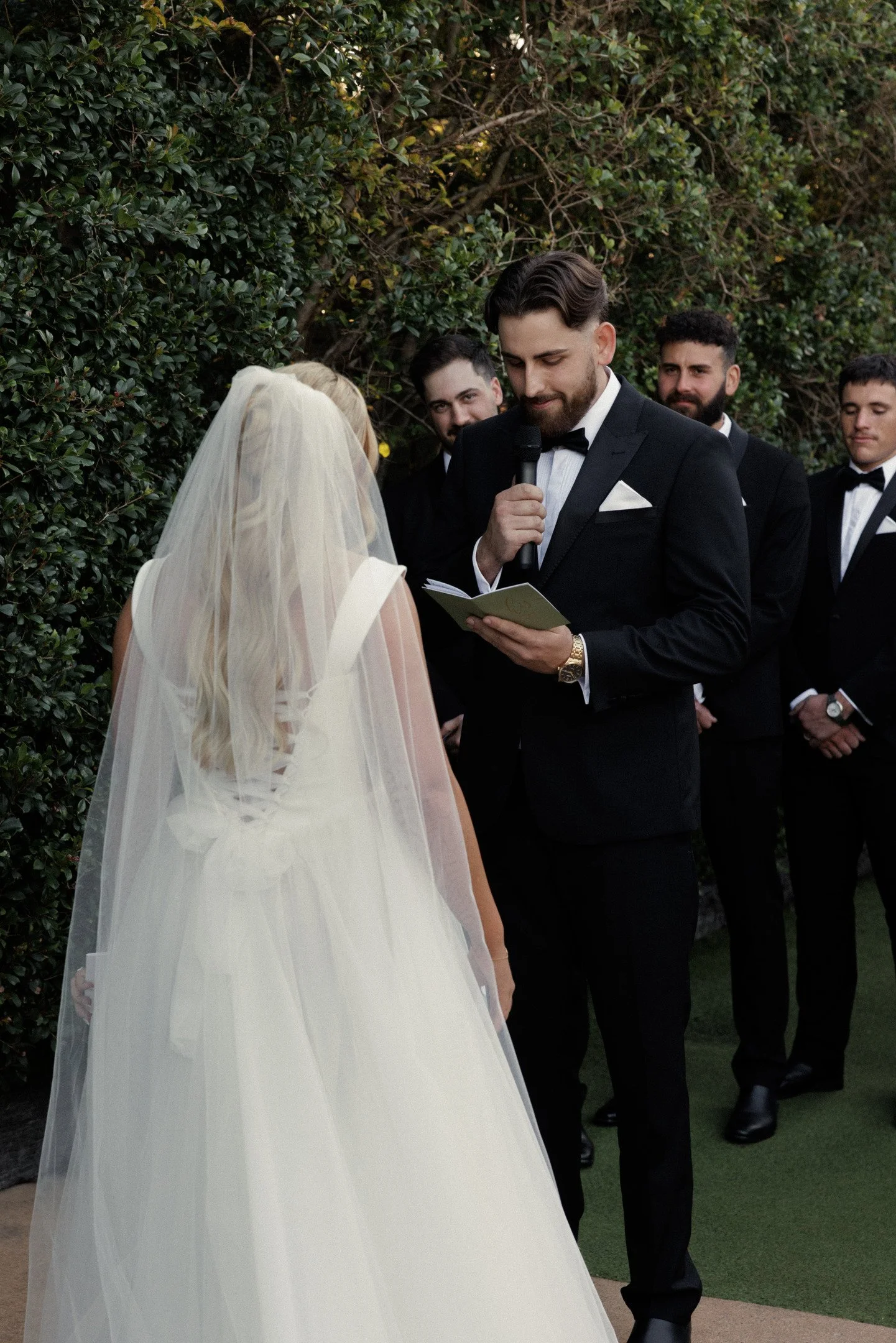 A wedding ceremony with a groom in a black tuxedo reading vows to a bride in a white wedding gown and veil, surrounded by groomsmen in tuxedos, outdoors with green bushes in the background.
