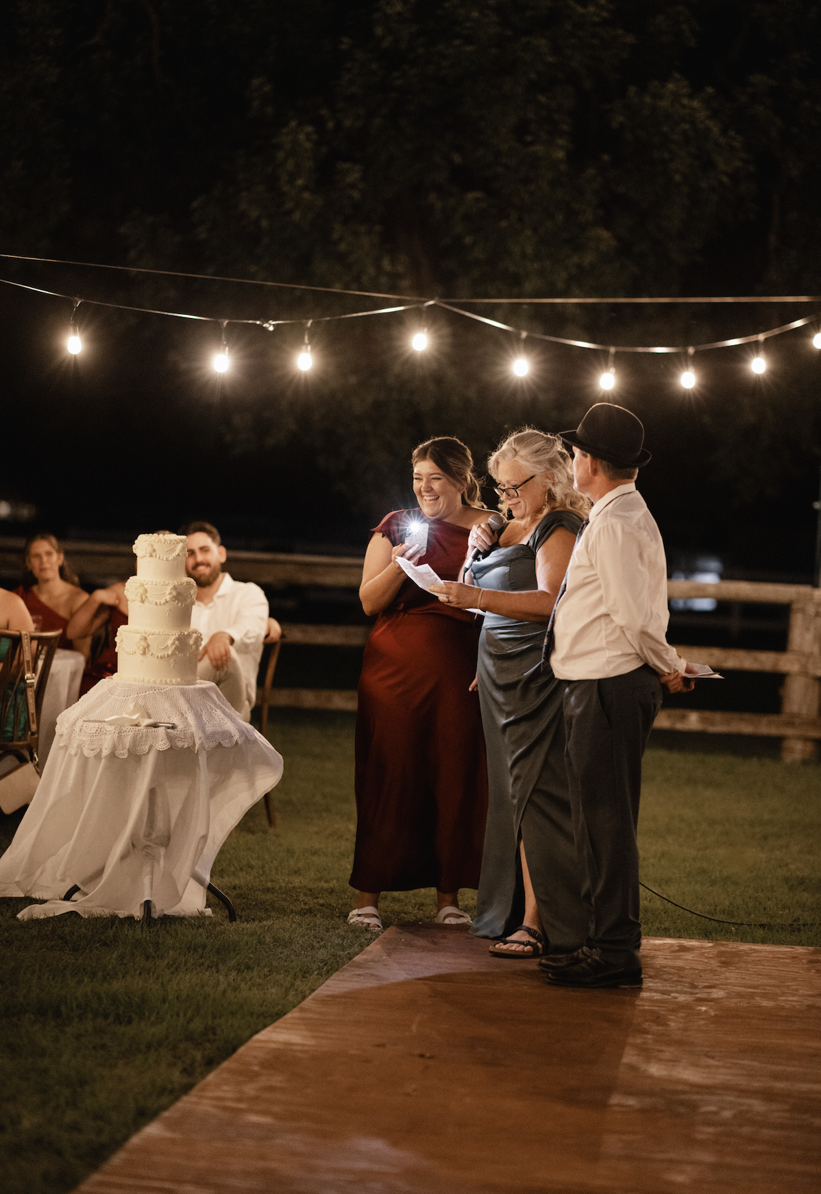 An outdoor wedding reception at night with string lights overhead. Three people are standing on a wooden dance floor, with one woman reading vows or speech into a microphone. A wedding cake is visible on a table to the left. Guests are seated at tabl