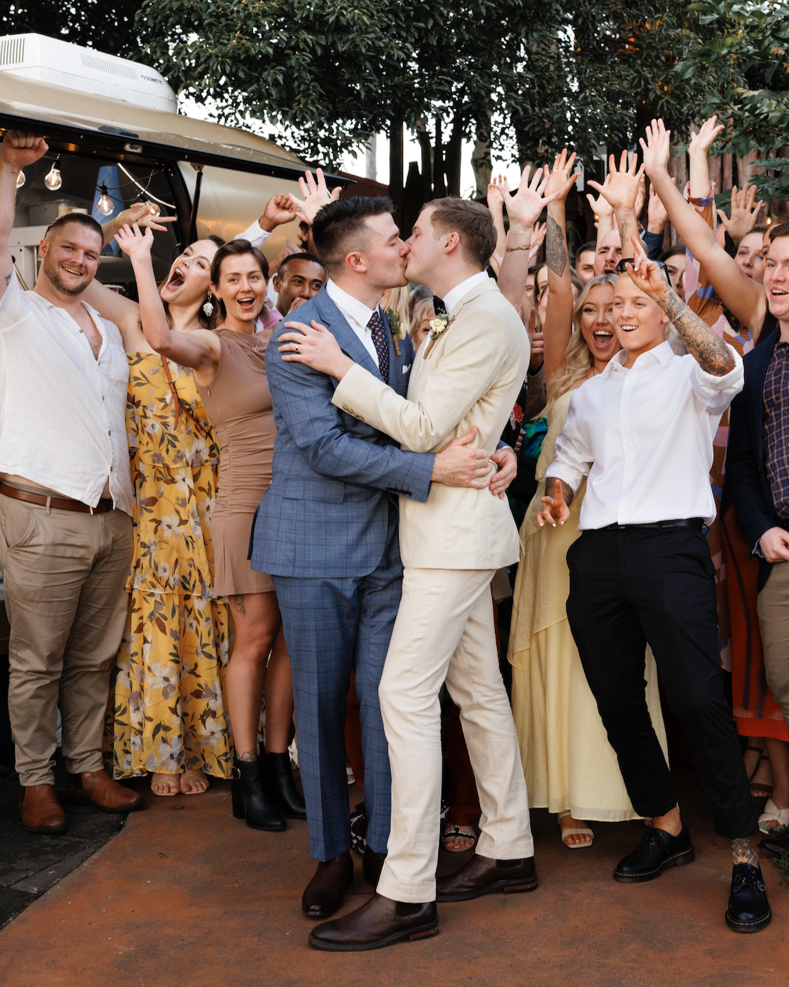 Two men kissing at a wedding reception, surrounded by a joyful crowd of people with raised hands, outdoors with trees and string lights in the background.