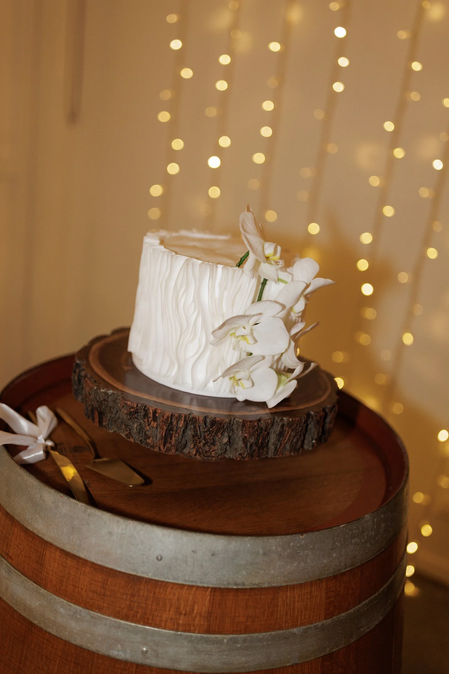 A wedding cake with white icing and orchids on top, placed on a round wooden slab. The background has warm lights creating a soft, festive atmosphere.