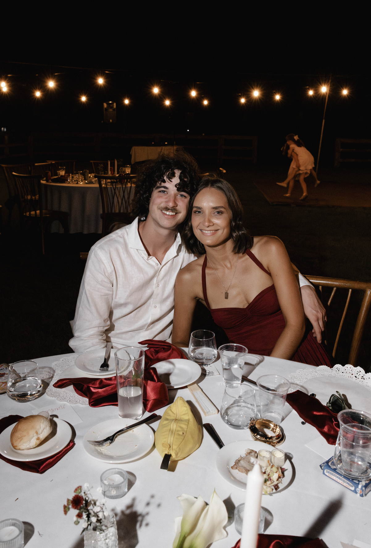 A smiling couple sitting at a dinner table during an outdoor nighttime event with string lights overhead, with children playing in the background.