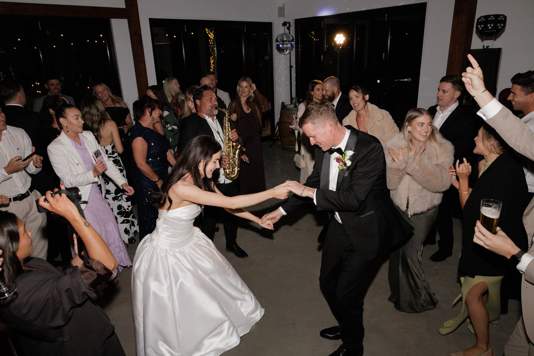 A wedding reception with guests dancing, including a bride and groom holding hands and dancing. A live band with a saxophone player is performing, and everyone appears to be enjoying the celebration.