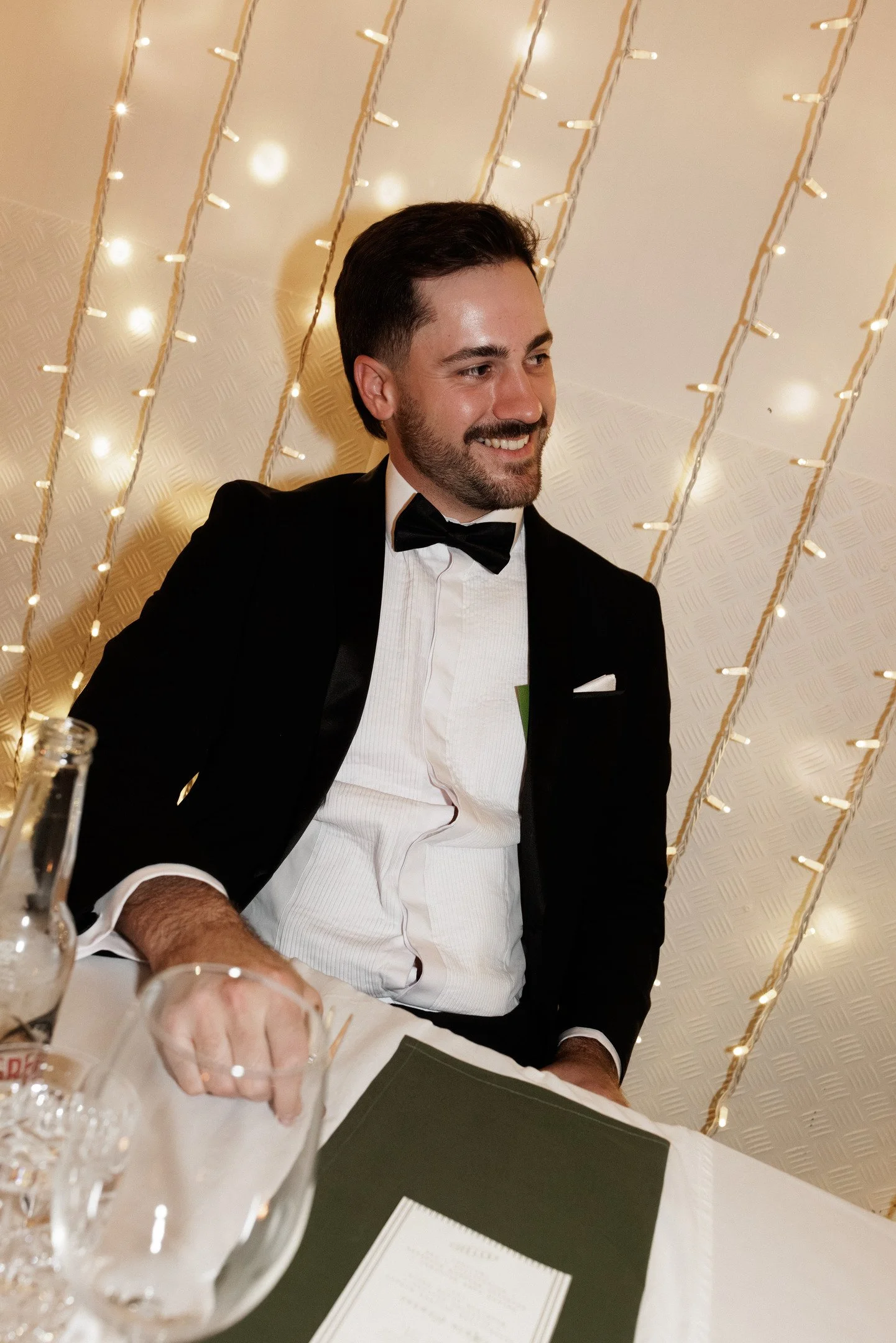A man with dark hair and a beard dressed in a tuxedo with a bow tie sitting at a table decorated with fairy lights in the background. He is smiling and appears to be at a formal event.