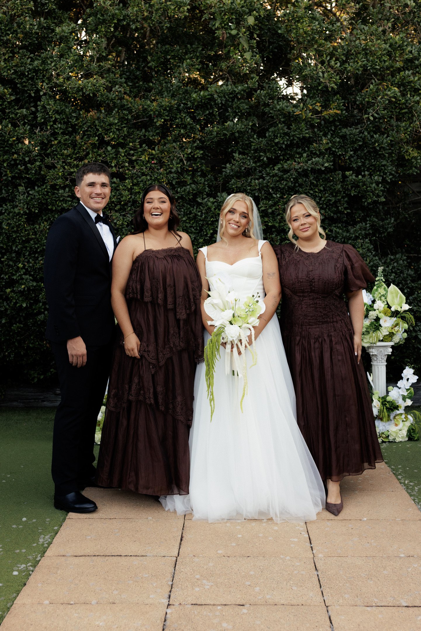 A group of four people, including a bride in a white wedding dress holding a bouquet of white flowers, and three women in dark brown dresses, standing outdoors in front of a dense green hedge. The groom is on the left in a black tuxedo with a bow tie