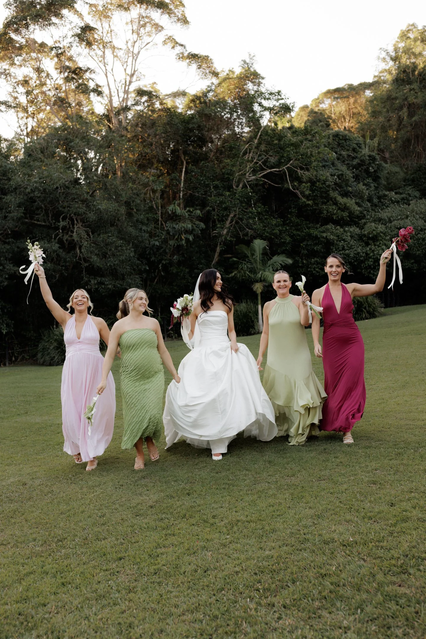 A bride and four bridesmaids walking on a grassy area outdoors, smiling and holding flowers, with trees in the background.