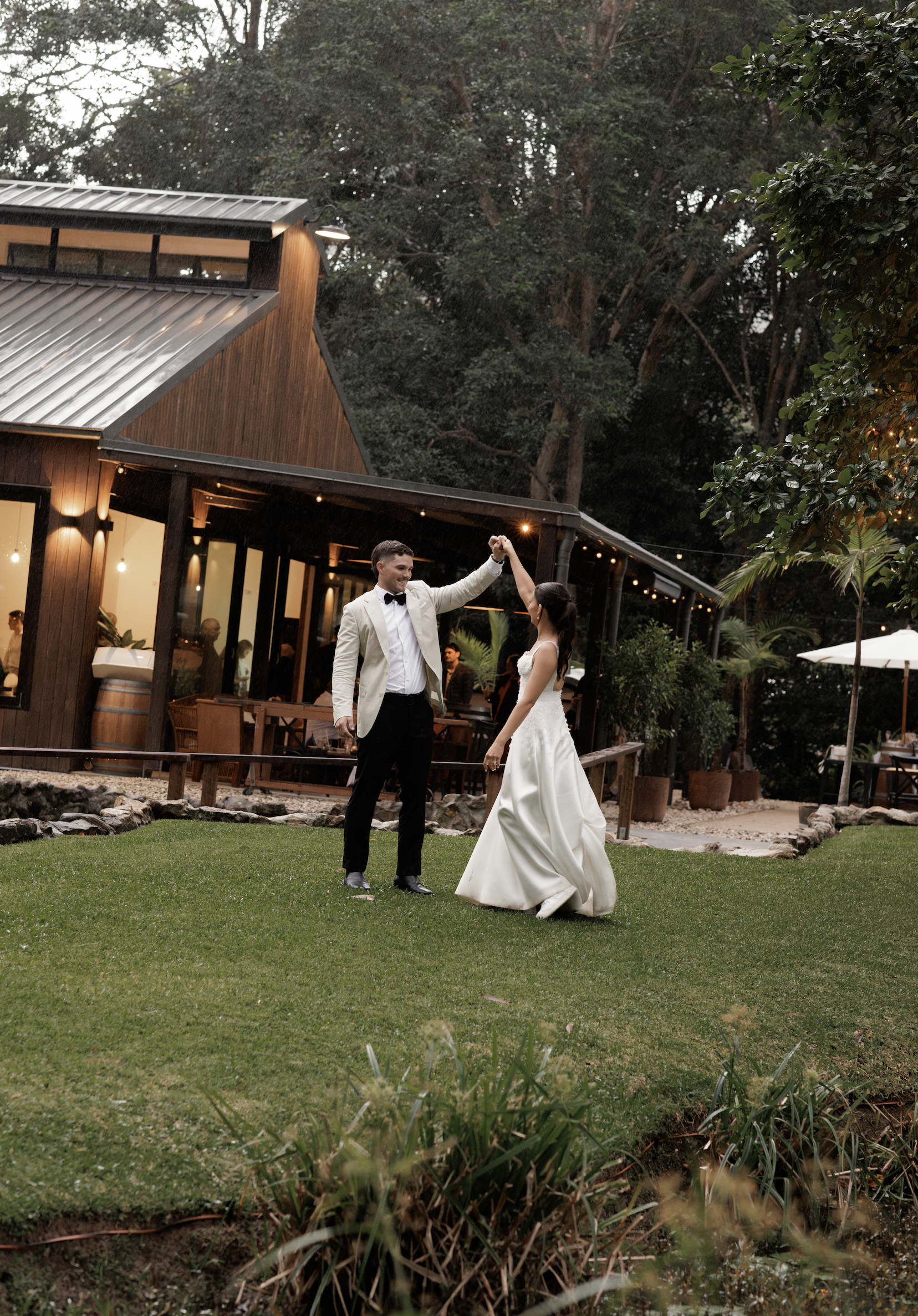 A couple dancing outdoors at night, with the man in a light-colored tuxedo and the woman in a white wedding dress, in front of a modern wooden building with string lights, trees, and bushes in the background.