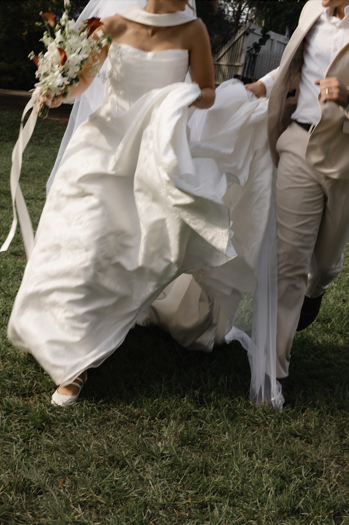 Bride and groom walking on grass, holding hands, wedding attire with the bride in a white gown and the groom in a light-colored suit.