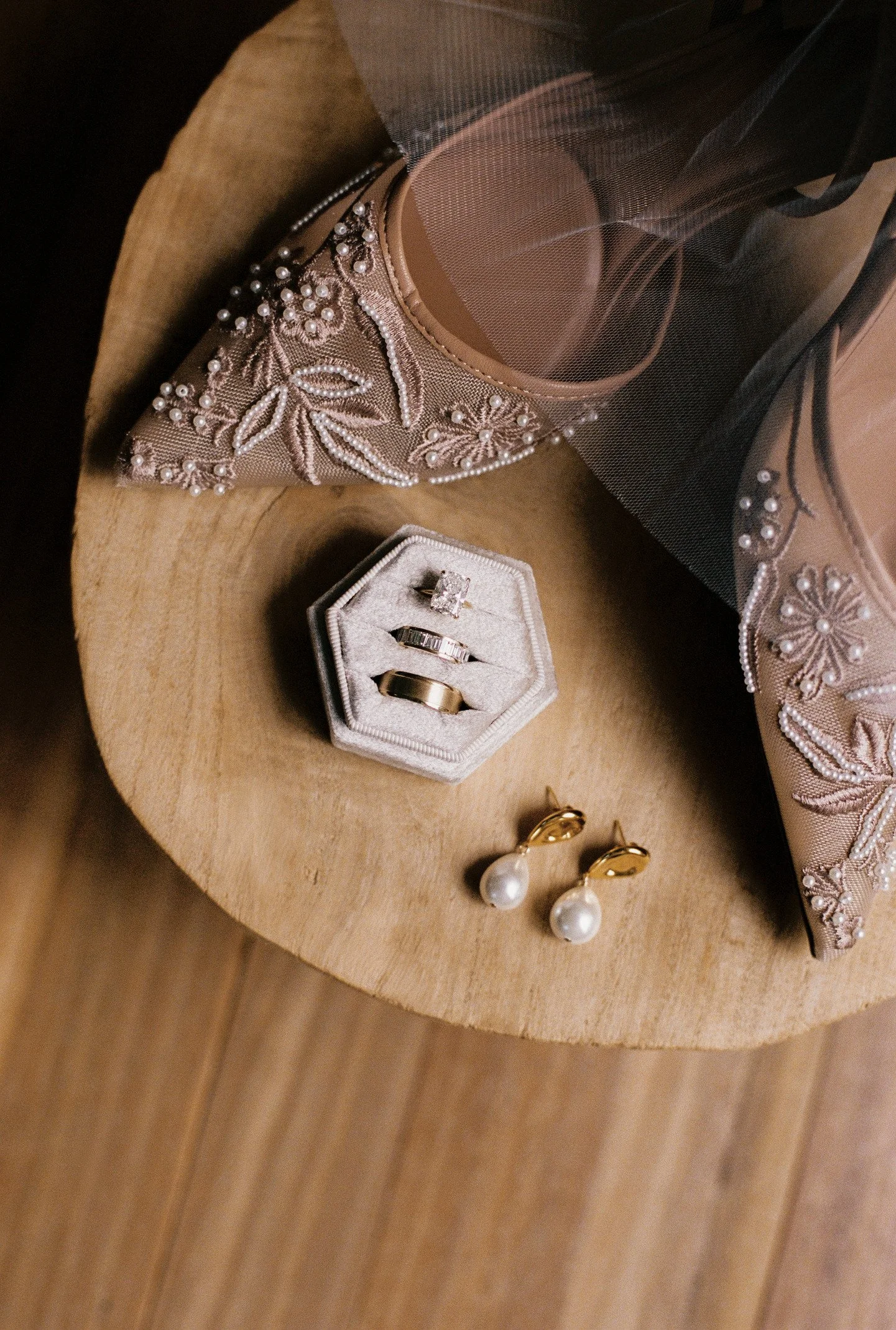 A beige and pink embroidered purse with floral design, placed on a wooden surface. Next to the purse, there is a small jewelry box with rings, and a pair of pearl earrings with gold hooks.