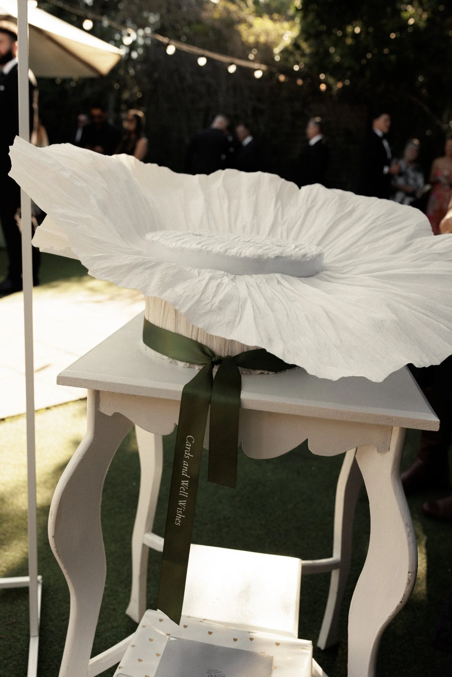 White bloom-shaped cake with green ribbon on a white table at an outdoor event with people in formal attire in the background.