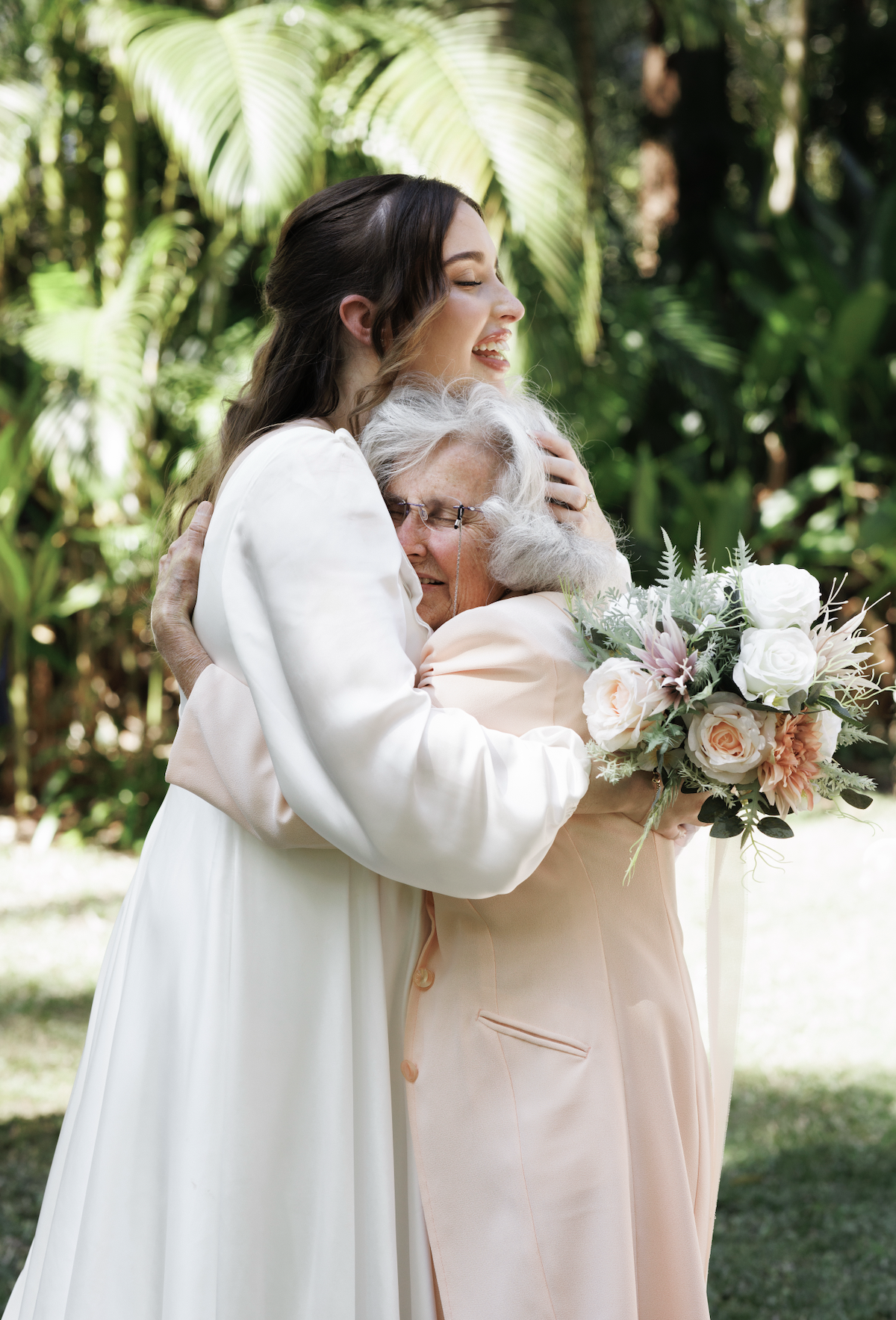 A young woman in a white dress is hugging an elderly woman with gray hair, who is holding a bouquet of flowers, in a lush green garden.