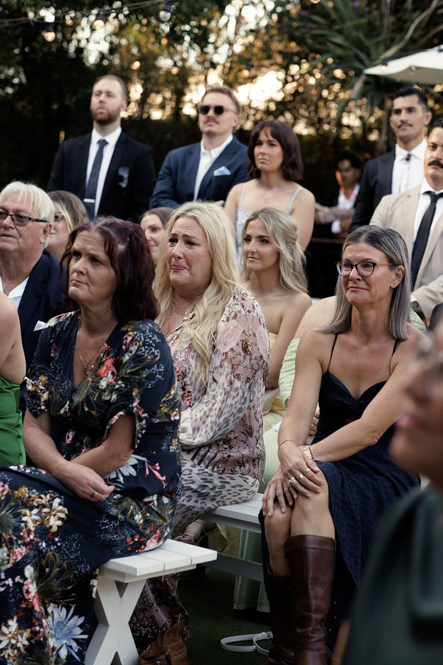 A group of people at an outdoor event, sitting and standing, dressed in semi-formal attire, with trees and sunset in the background.