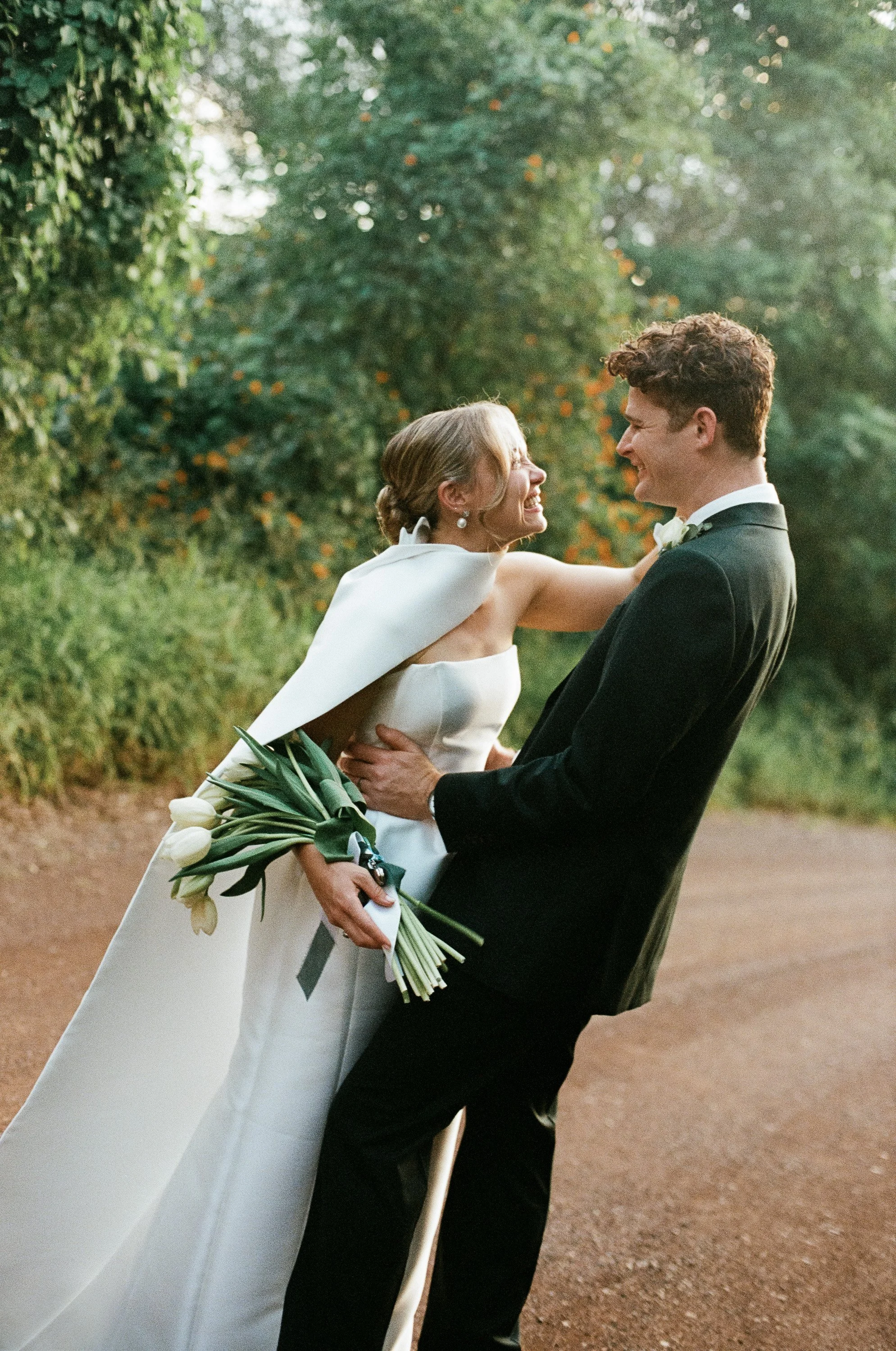 A bride and groom sharing a joyful embrace on a forest path during their wedding, with the bride holding a bouquet of white tulips.