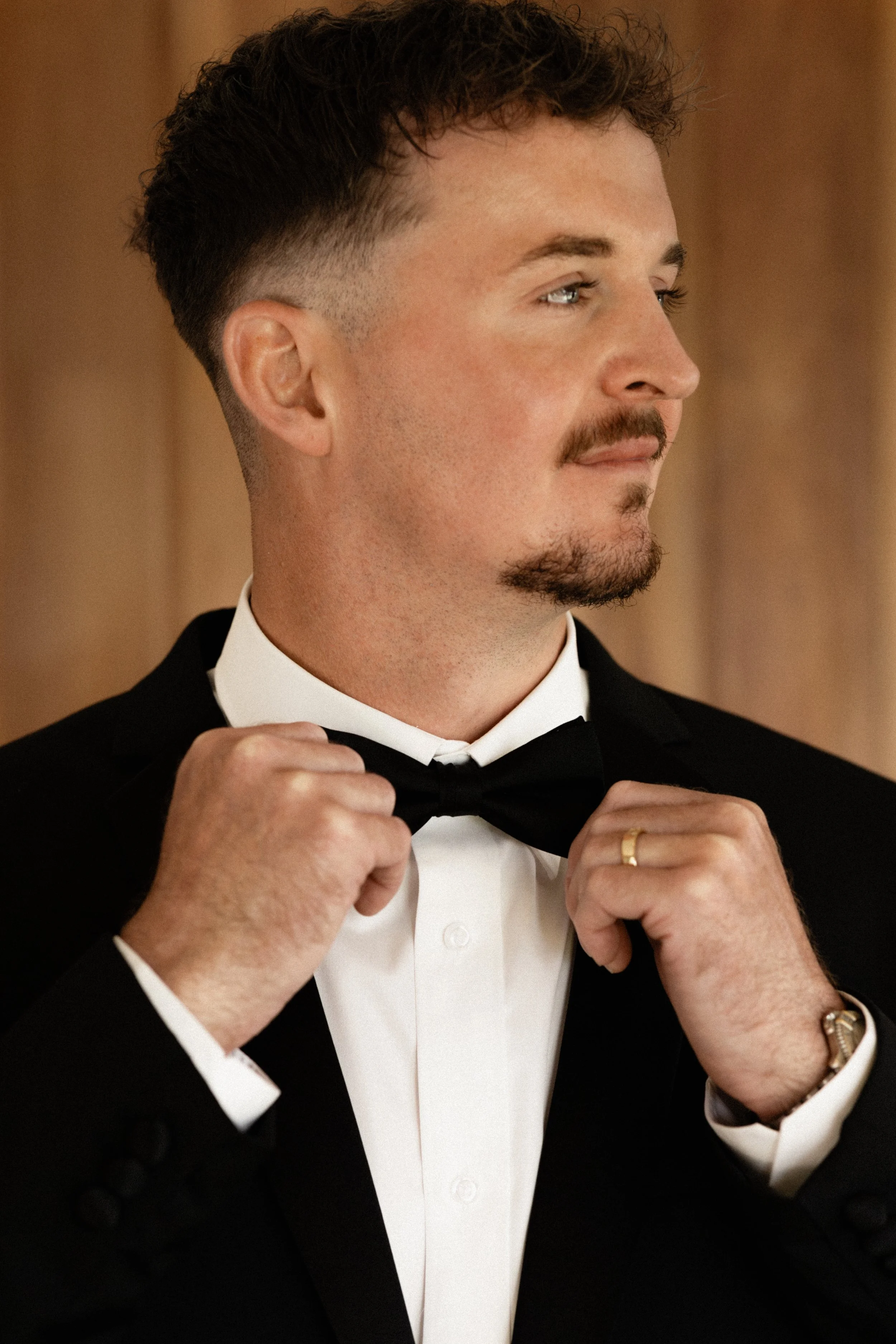 A man dressed in a tuxedo adjusting a black bow tie with a focused expression, standing in front of a blurred warm-colored background.