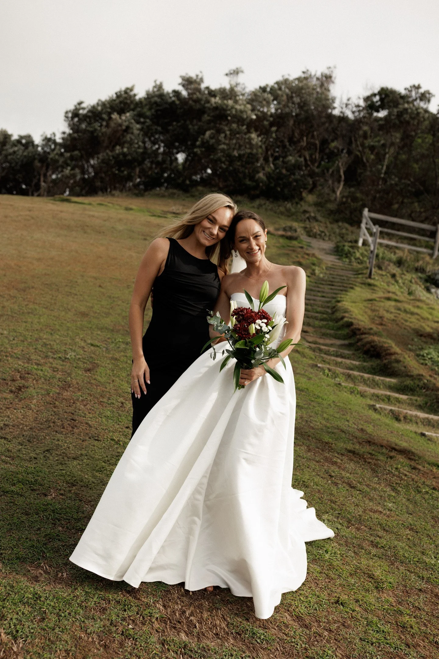 Two women are smiling and posing outdoors on a grassy hill, with one wearing a black dress and the other in a white wedding gown holding a bouquet of flowers.