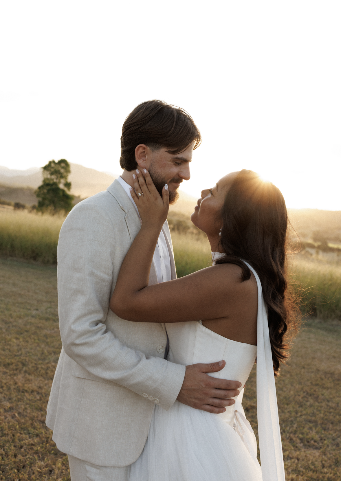 A couple embraces outdoors during sunset, gazing into each other's eyes, with hills and a few trees in the background.