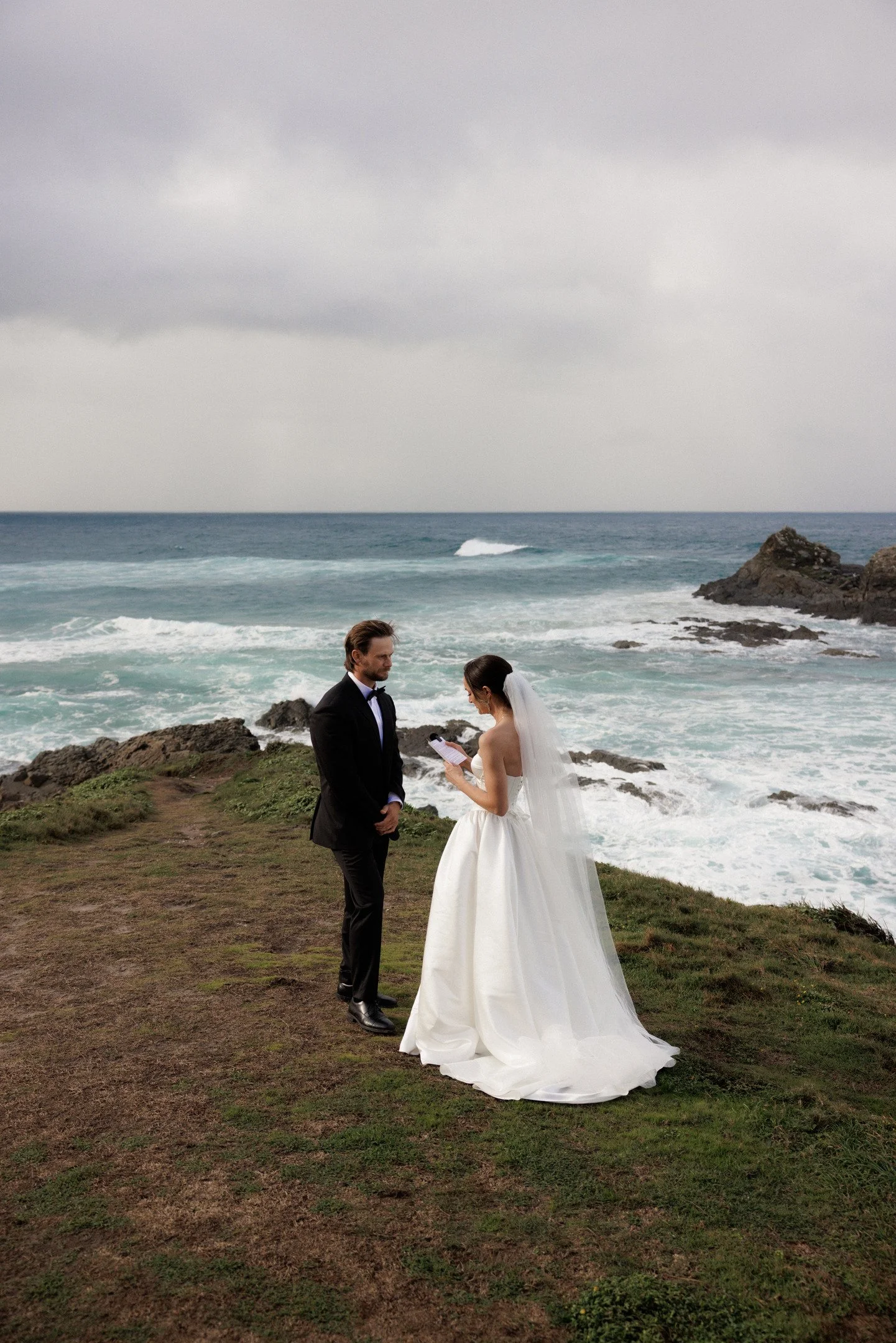 A couple in wedding attire standing on a grassy area near the ocean, with the groom in a black tuxedo and the bride in a white wedding dress, exchanging vows facing each other against a cloudy sky and rocky shoreline.
