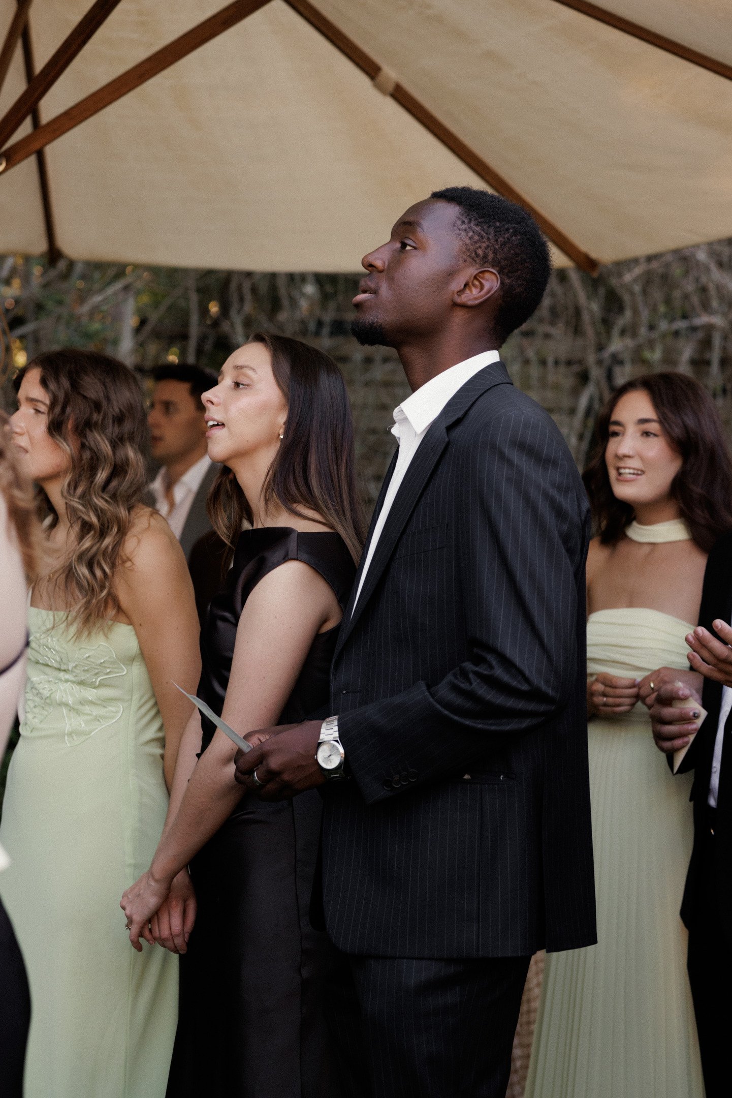 A group of people standing in a line at an outdoor event, dressed in formal attire. The focus is on a young Black man in a dark pinstripe suit holding a piece of paper, with women around him wearing pastel-colored dresses.