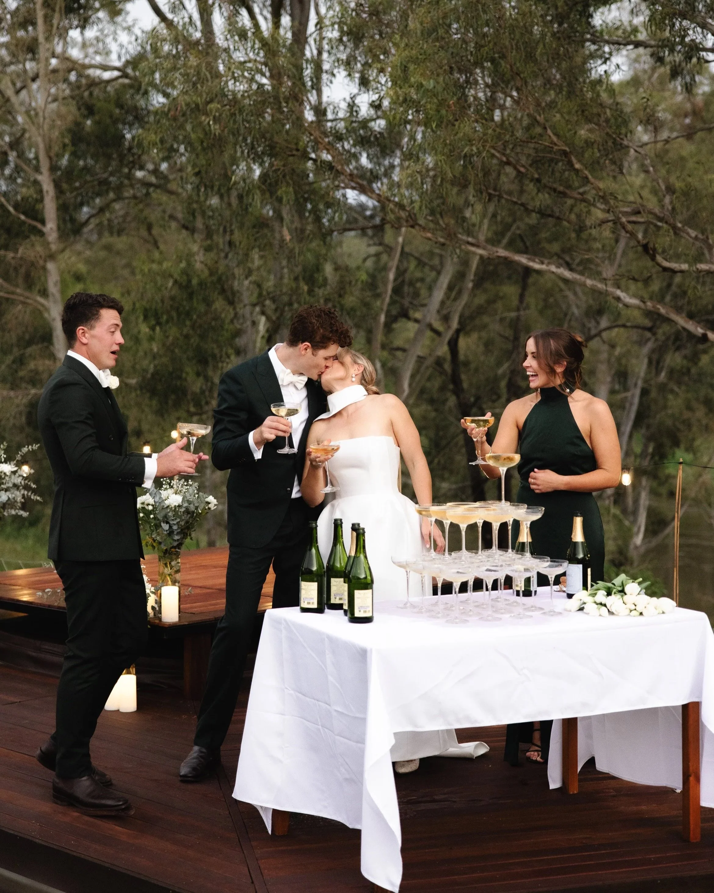 A wedding reception outside with four people, two men and two women, dressed in formal attire. The bride and groom are sharing a kiss while holding champagne glasses. A champagne tower is on a table in front of them, along with bottles of champagne and white floral arrangements. Everyone is smiling and celebrating.