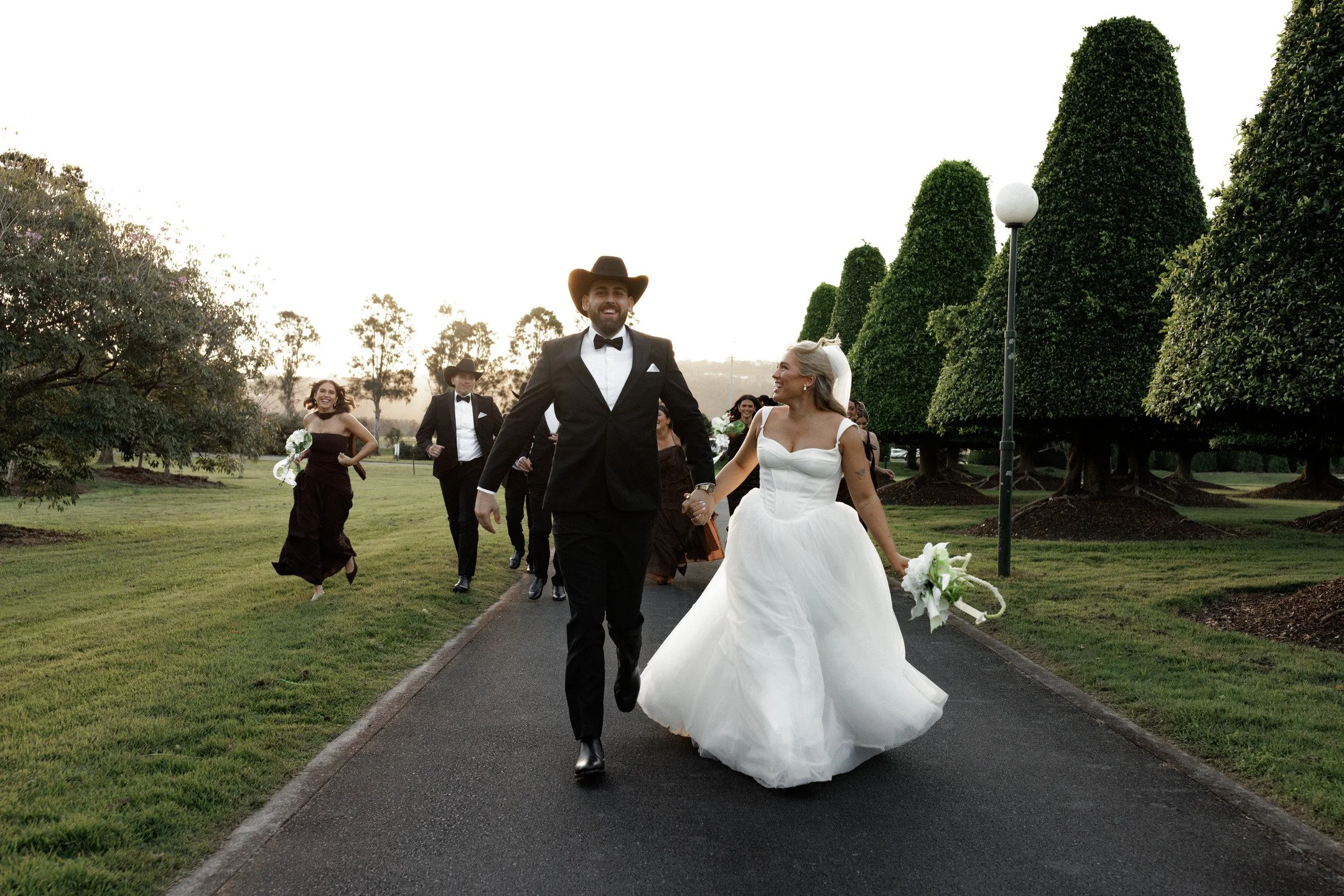 A group of people, including a bride in a white wedding gown and a groom in a black tuxedo, walking hand in hand on a pathway in a park or garden during sunset. The bride is holding a bouquet, and there are tall, trimmed trees and a lamp post in the 