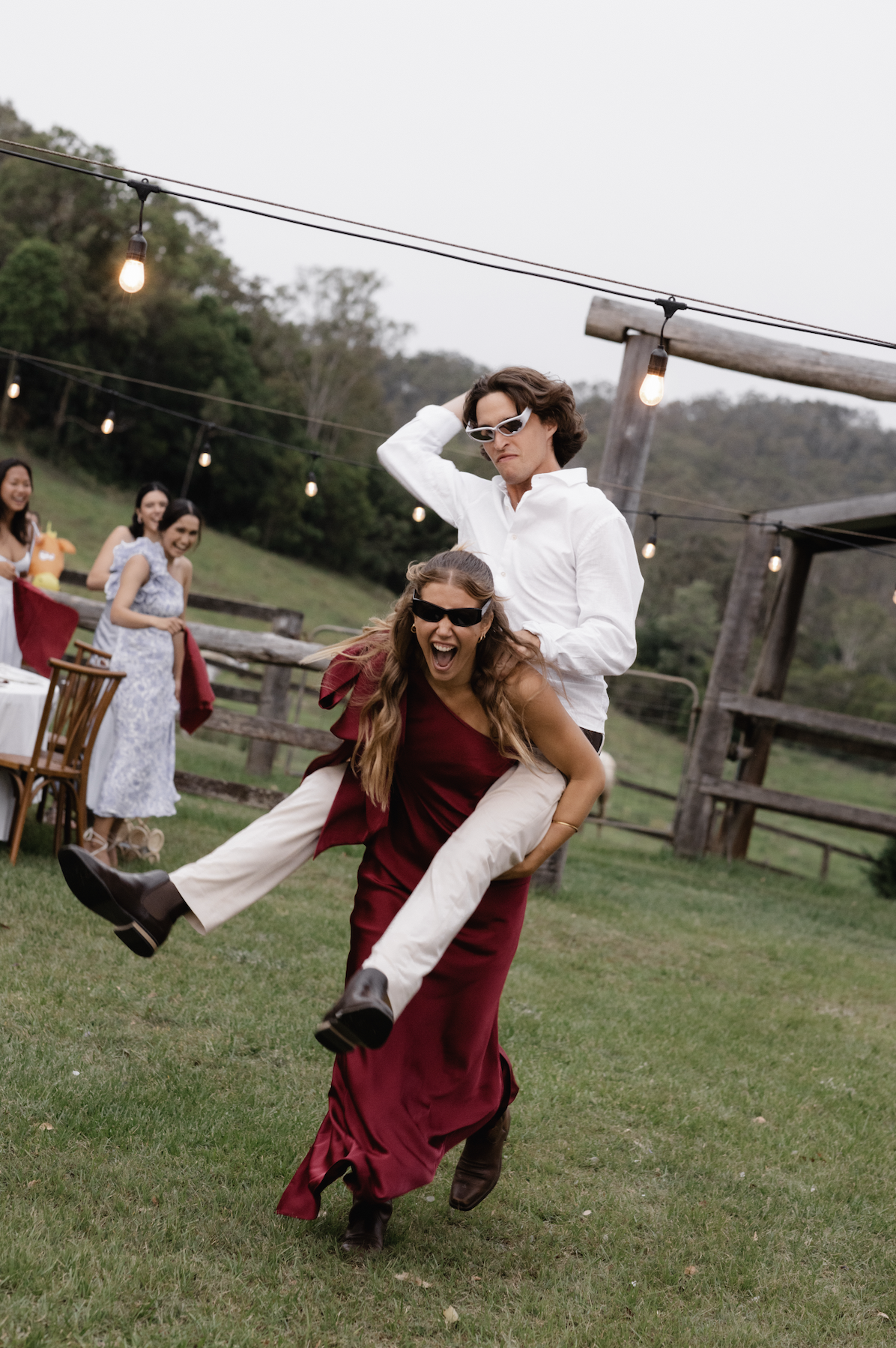 People dancing and celebratin outdoor event on grassy field with string lights and trees in background, woman in red dress being carried on shoulders