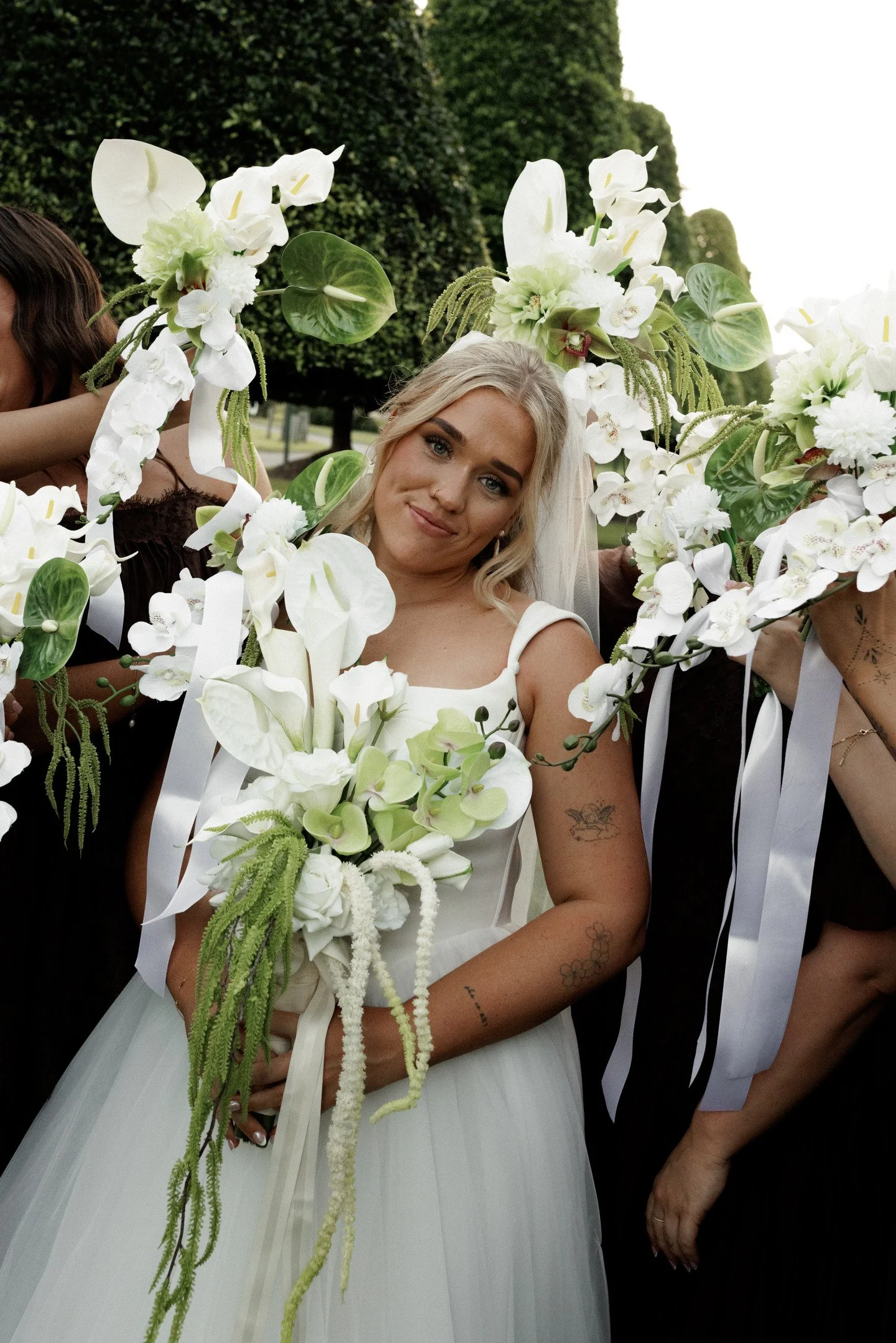 A woman in a white dress holding a large bouquet of white and green flowers, surrounded by other women holding similar bouquets, outdoor scene with trees and overcast sky.