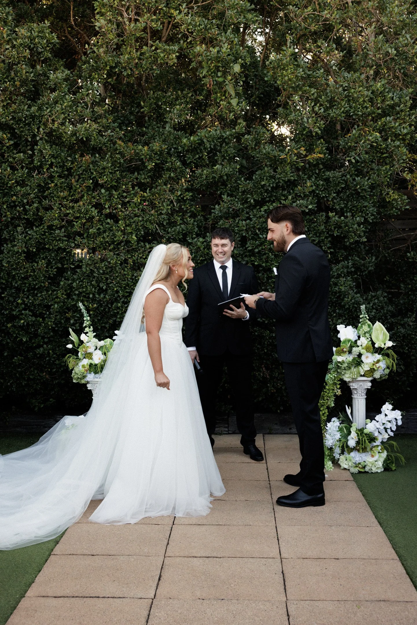 A bride and groom stand facing each other during their wedding ceremony, with a male officiant standing between them, in front of a green leafy hedge. The bride wears a white wedding gown with a long train and veil, and the groom wears a black tuxedo